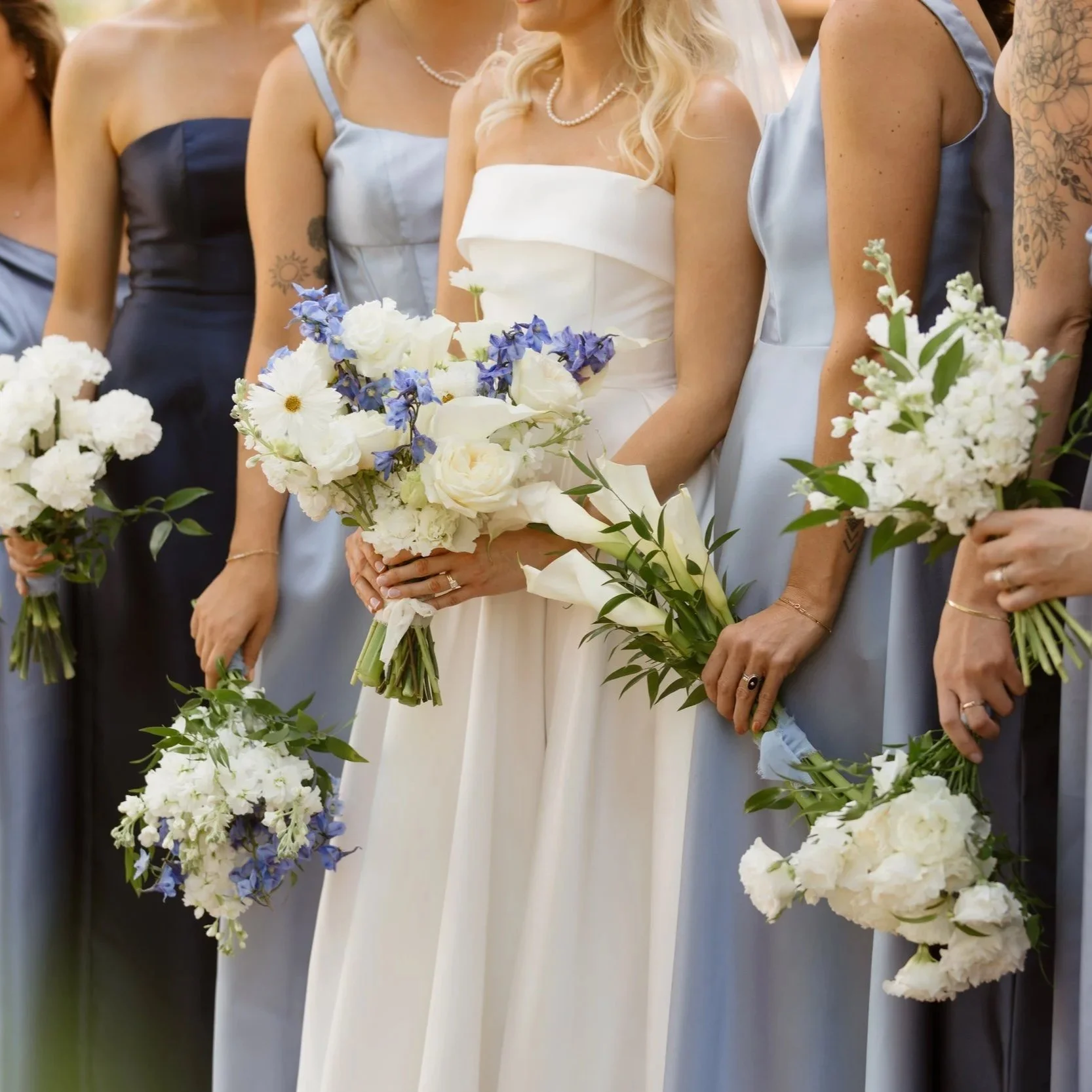 Bride in a white wedding dress holding a bouquet of white and purple flowers, standing among bridesmaids in blue dresses holding similar bouquets.