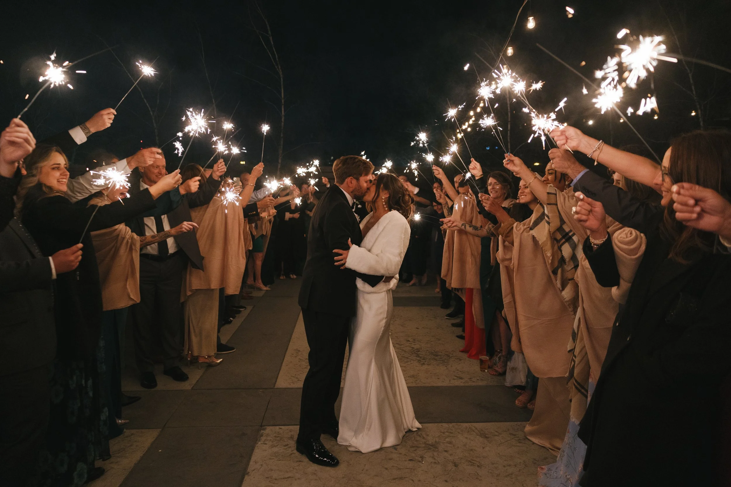 A wedding celebration at night with a bride and groom kissing in the center, surrounded by guests holding sparklers forming a tunnel for them.
