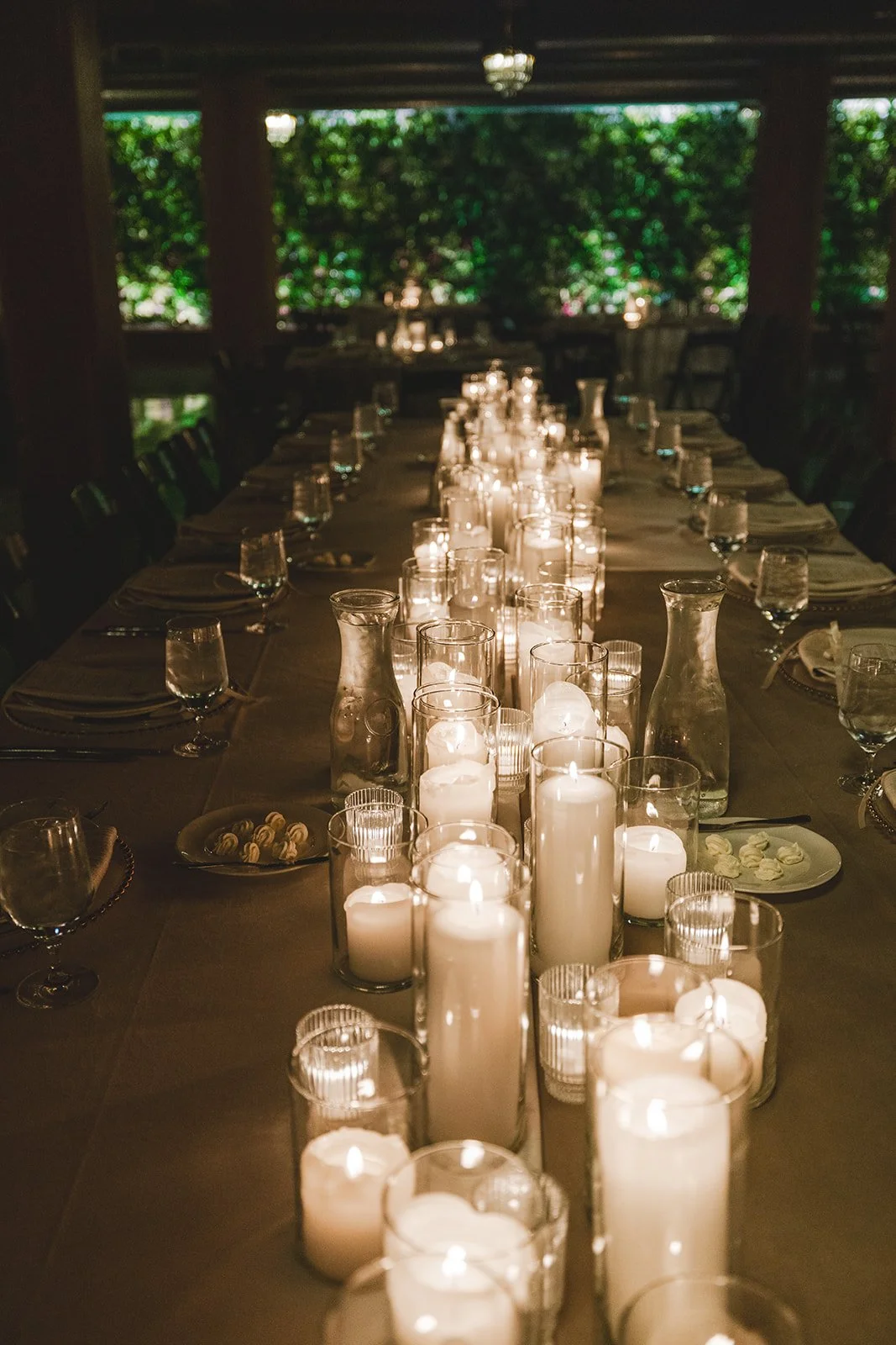 A long dining table decorated with numerous candles in glass holders and carafes, set for a celebration or elegant dinner in a dimly lit room with large windows and green foliage outside.