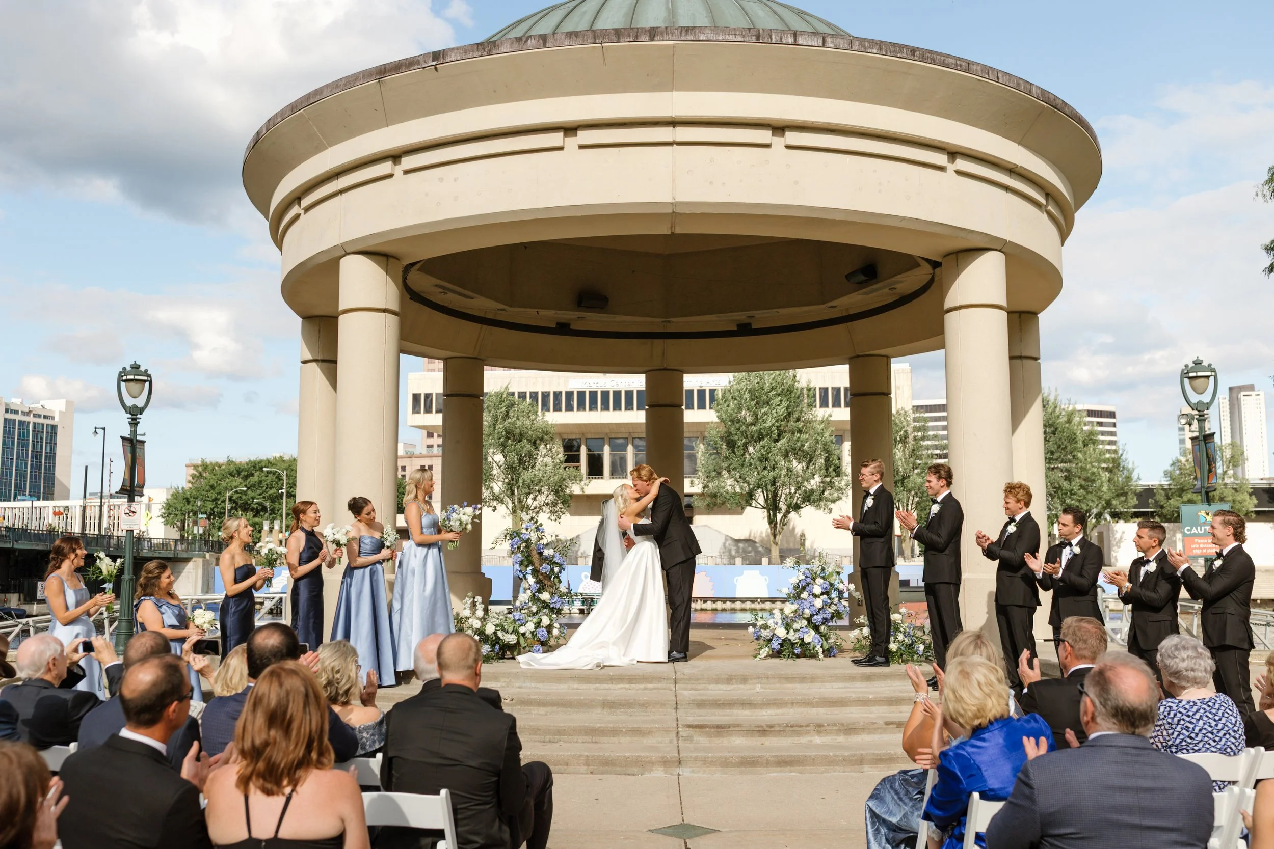 A wedding ceremony taking place outdoors in a round pavilion with tall columns. The bride and groom are kissing under an arch decorated with flowers, while guests are seated and standing around, watching and taking photos. The sky is partly cloudy.