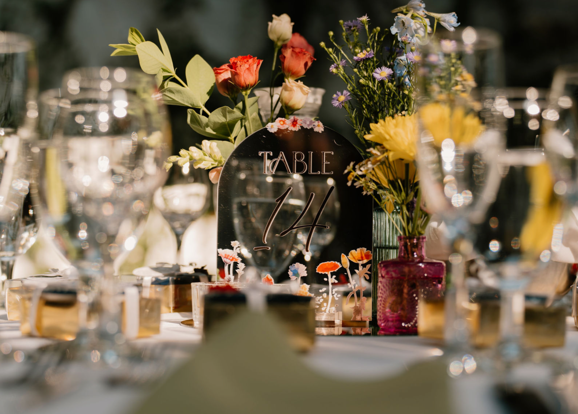 A decorated table with floral arrangements in colorful vases and a sign that says 'Table 14' amidst glassware and candles, set for a celebration or event.