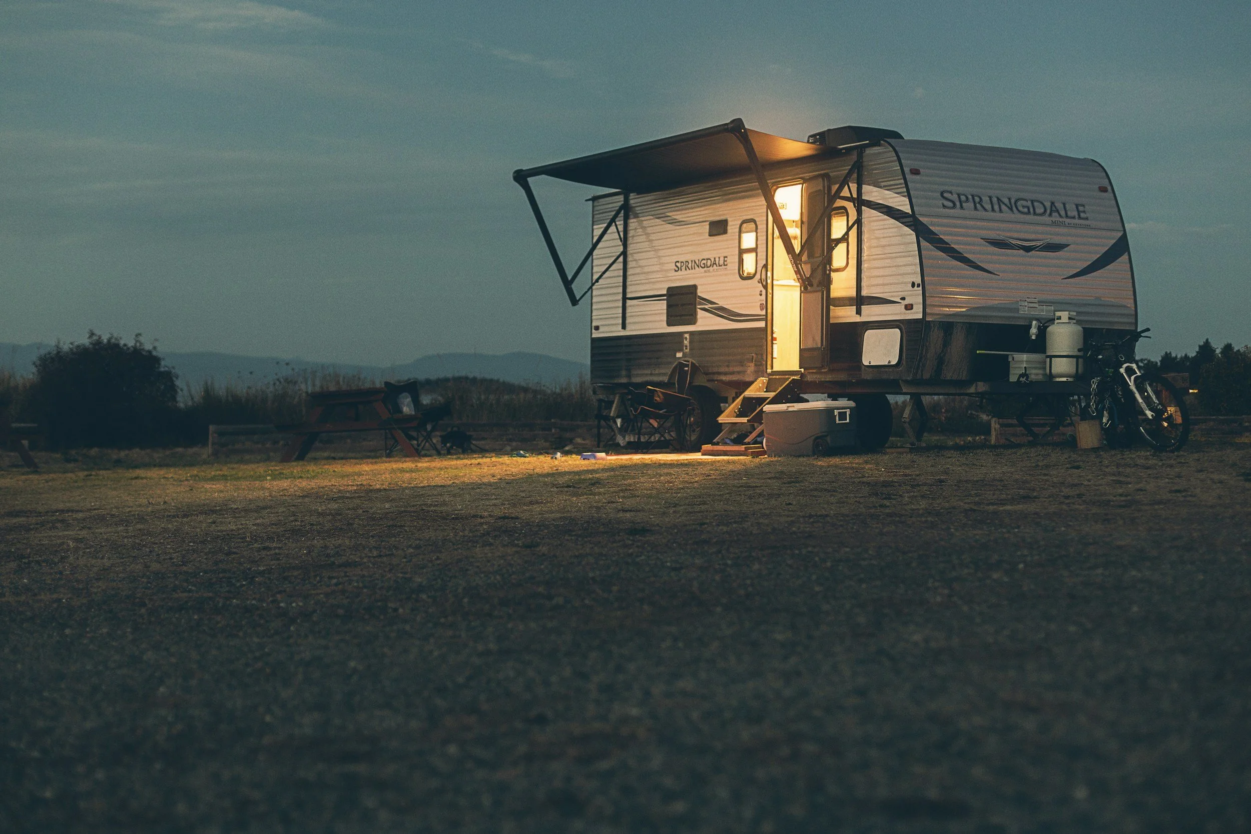A camping trailer with lights on in a rural outdoor setting at dusk, with outdoor chairs, a cooler, a bicycle, and propane tanks nearby.