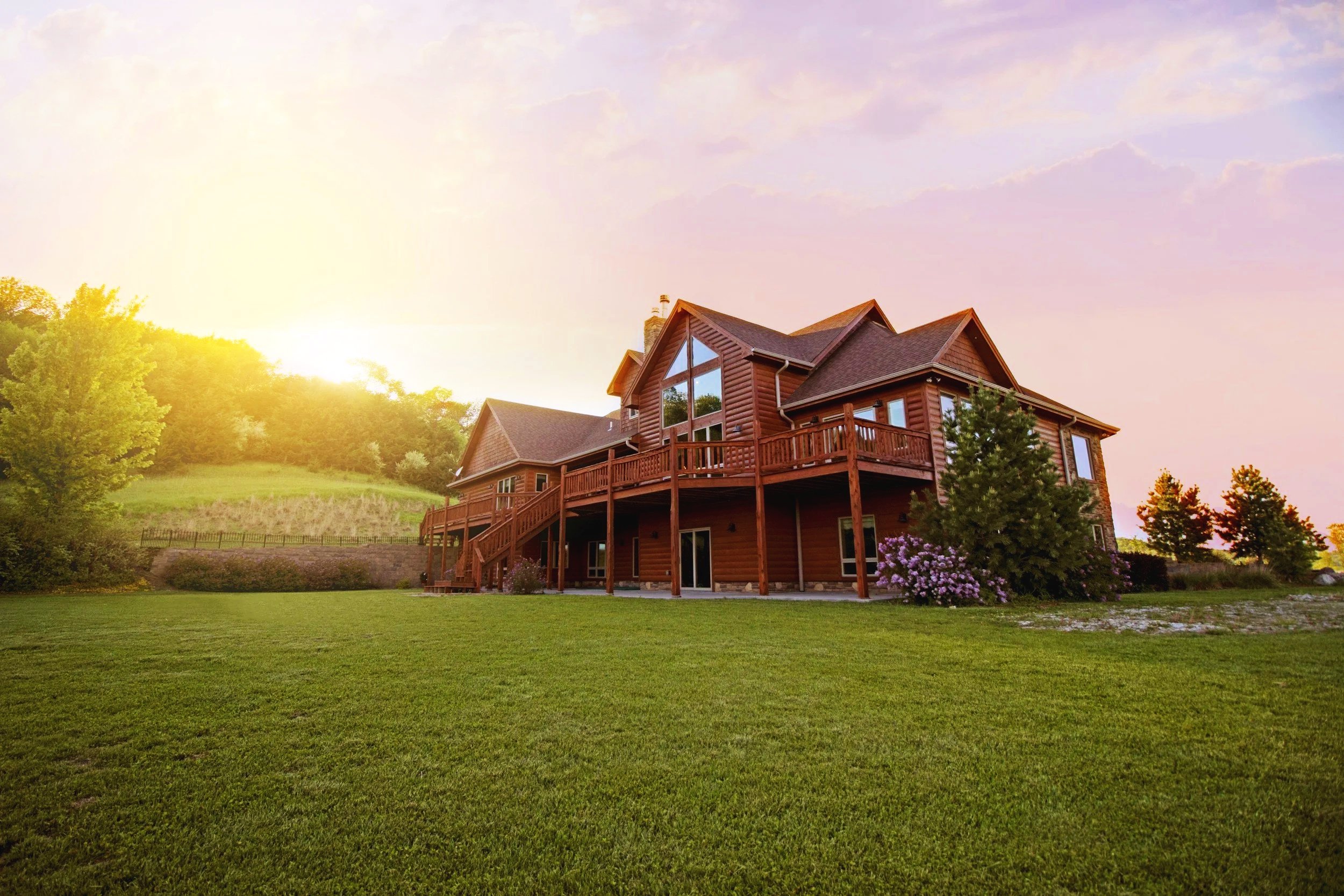 A wooden house with large windows, surrounded by green grass and trees, under a partly cloudy sky at sunset.
