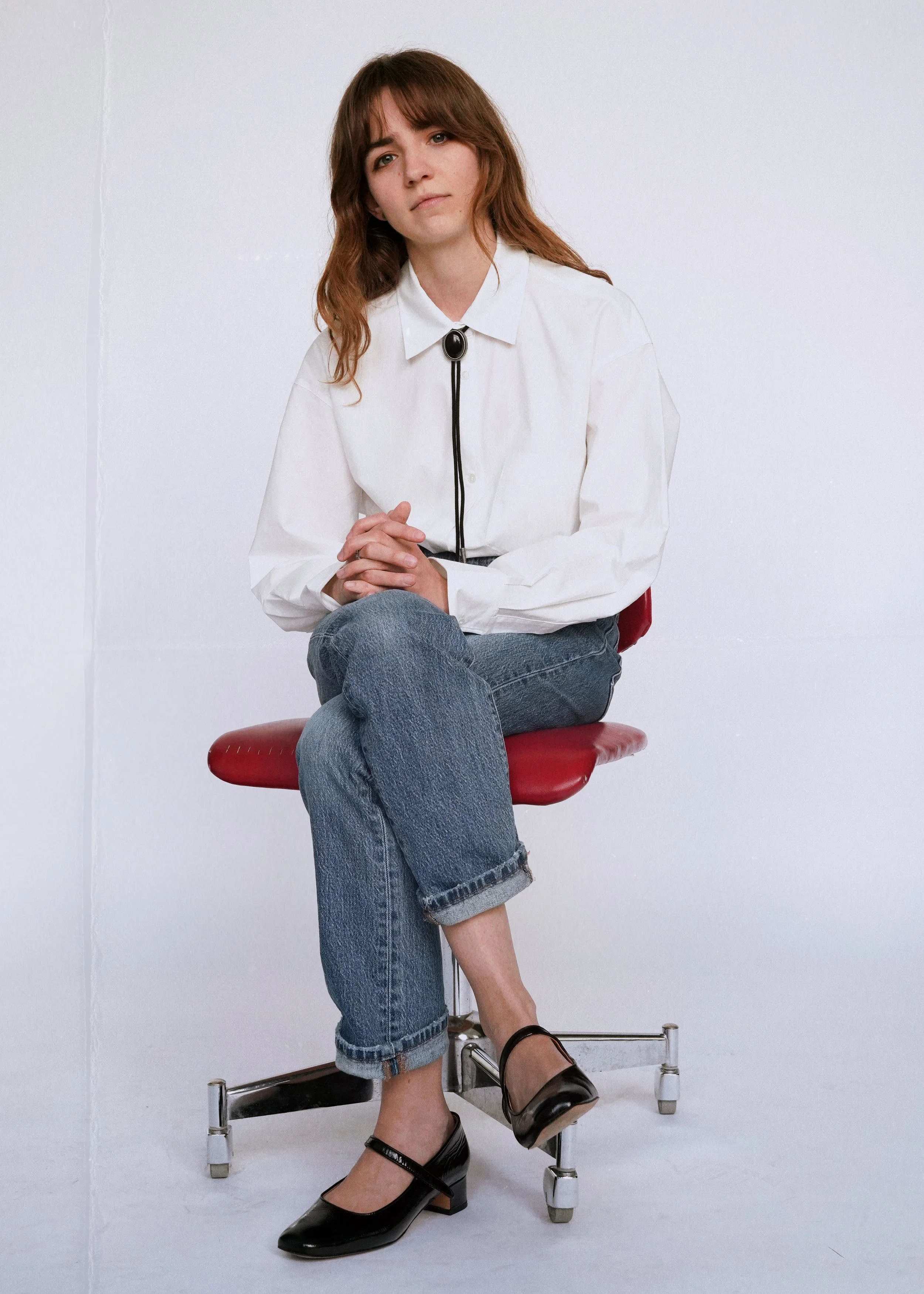 A woman with brown hair and a white shirt sitting on a red chair against a plain white background, with her hands clasped and looking slightly confused.