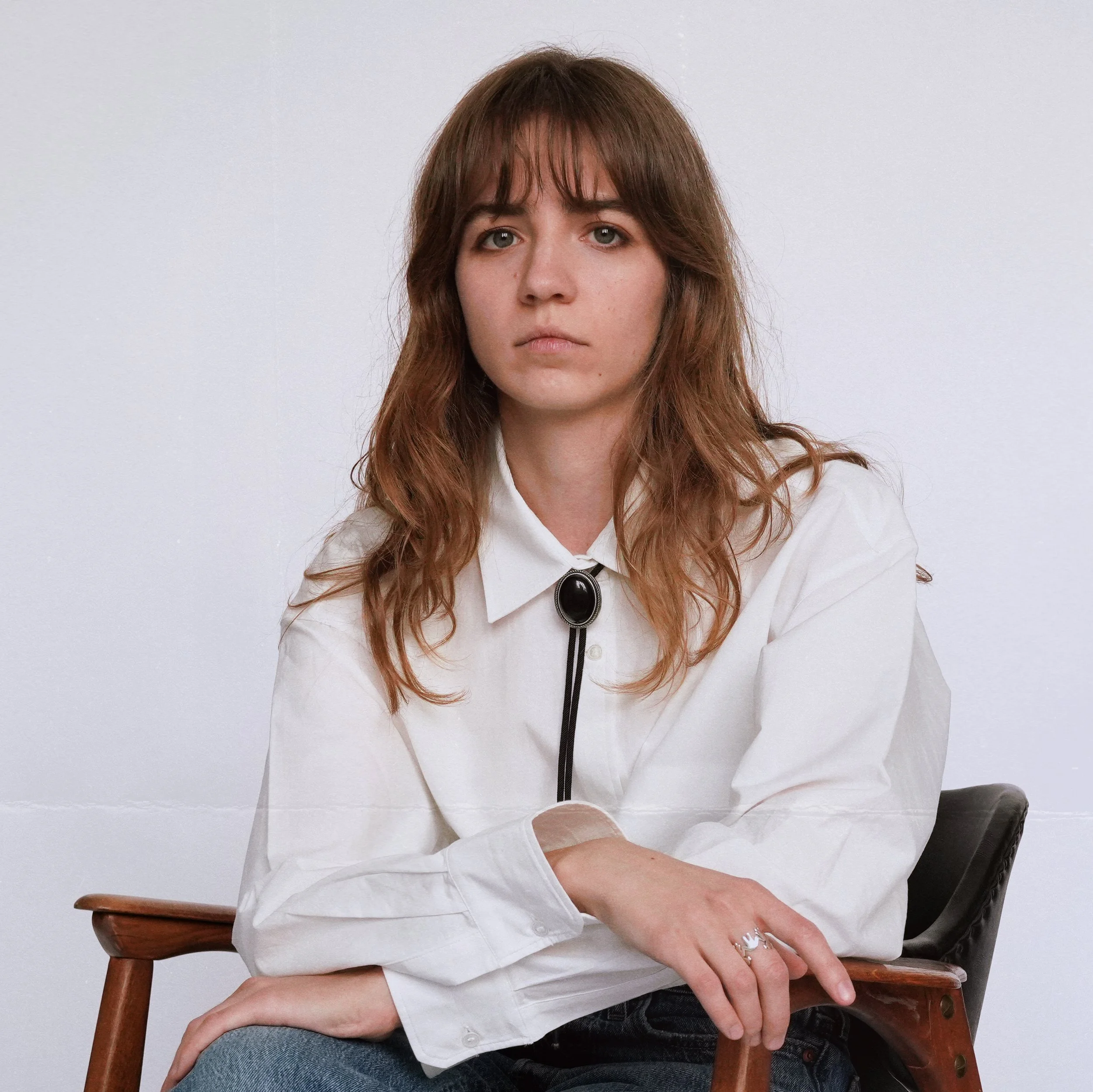 Portrait of a young woman with brown wavy hair, wearing a white button-up shirt with a black bolo tie, sitting on a wooden chair against a plain white background.
