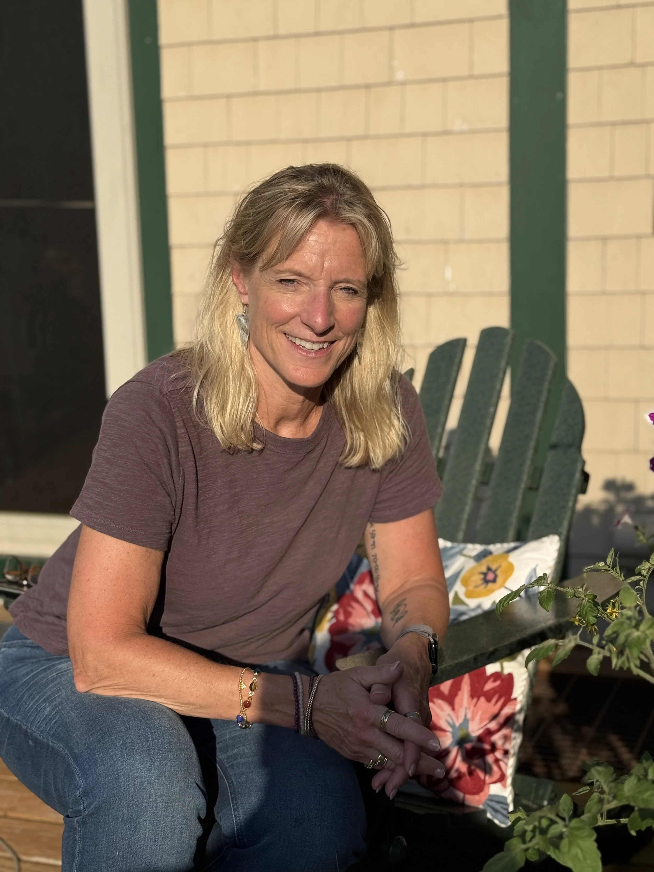 A smiling woman with long blonde hair, wearing a maroon T-shirt, blue jeans, and jewelry, is sitting outdoors at night with a patio background that includes green chairs and a yellow wall.
