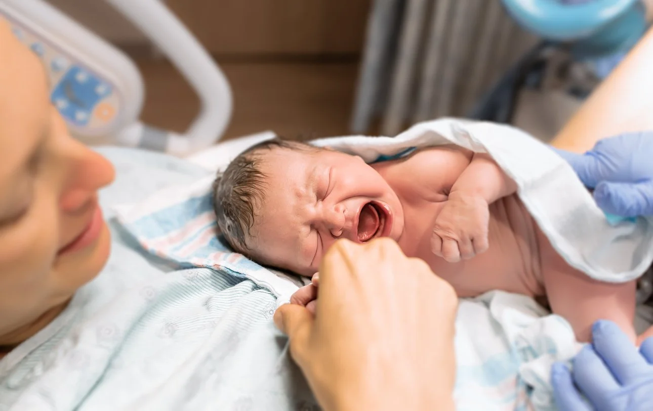 Newborn baby crying while being held by a woman, possibly a mother, in a hospital setting with medical staff assisting.