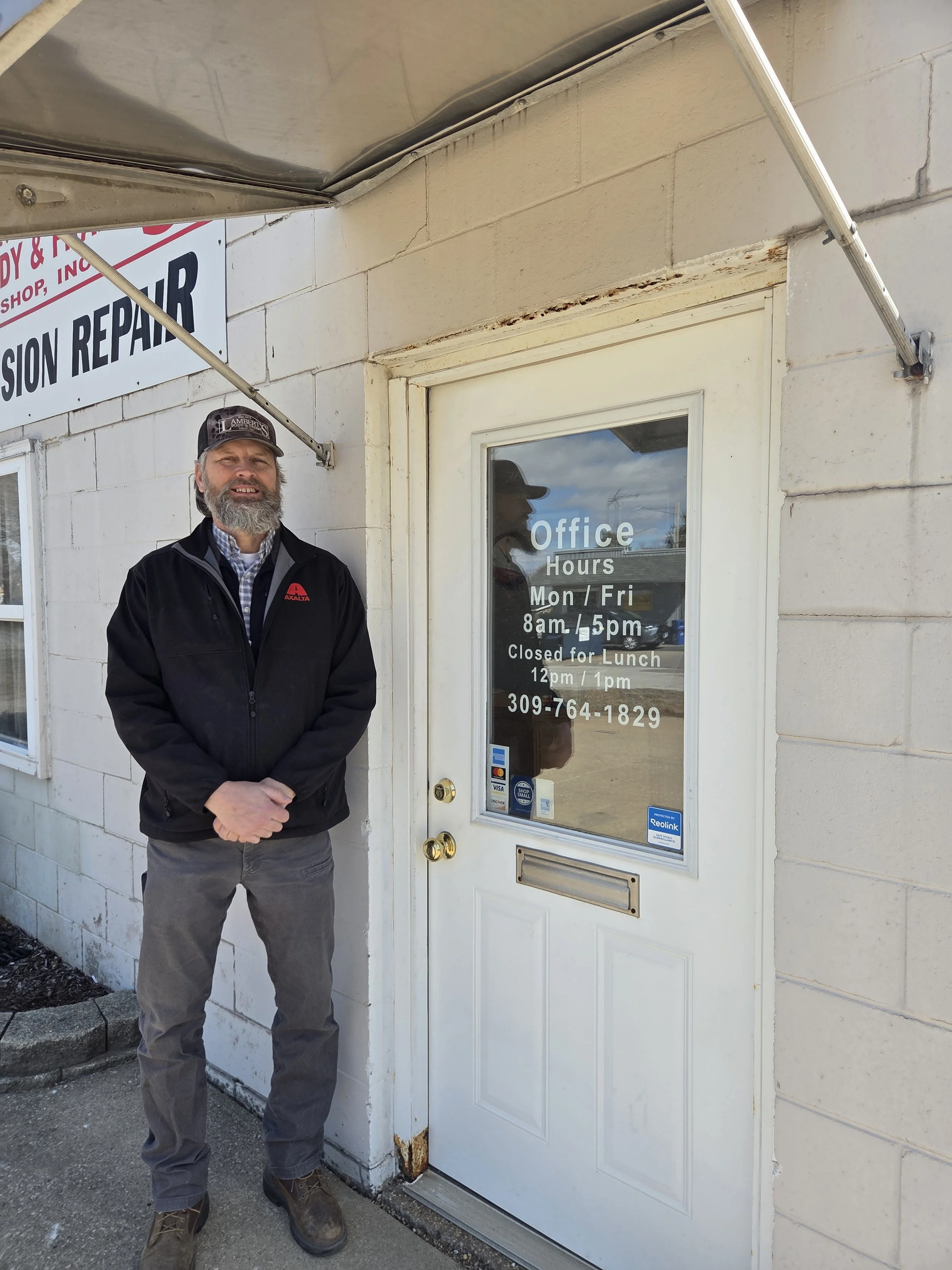 A man with a beard standing outside a building, next to a door with office hours written on it. The office is open Monday through Friday from 8am to 5pm, closed for lunch from 12pm to 1pm, with a phone number listed.