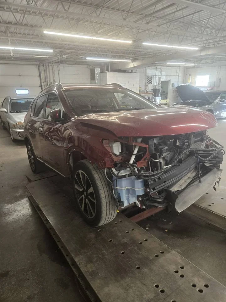 A red SUV with front-end damage in an indoor repair shop, missing its front bumper, headlights, and grille, with some internal components exposed.