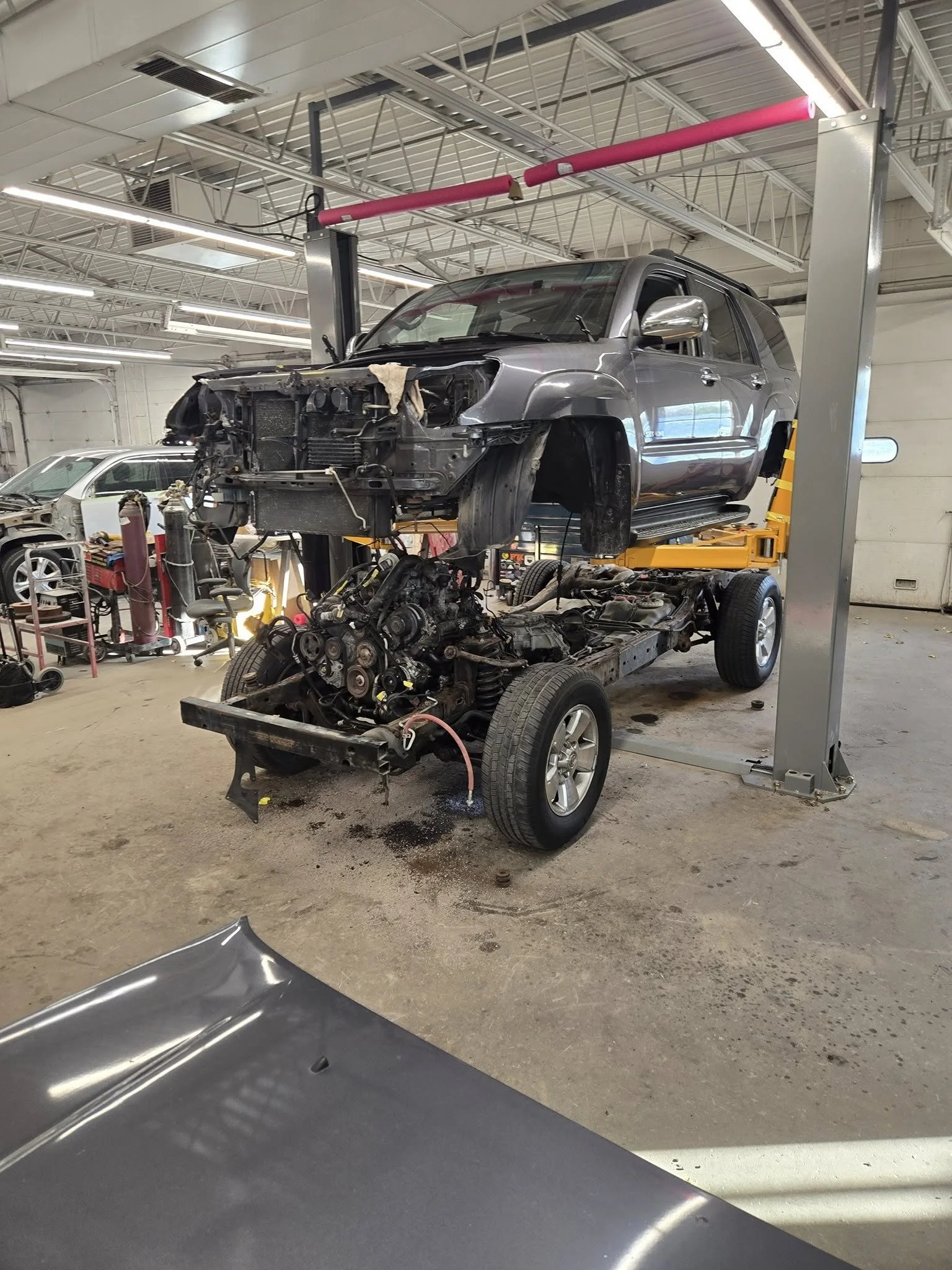 The image shows a partially disassembled SUV on a hydraulic lift in an auto repair shop. The front of the SUV is missing its engine and other components, exposing the engine bay. The body of the SUV is silver, and the shop has various tools and parts