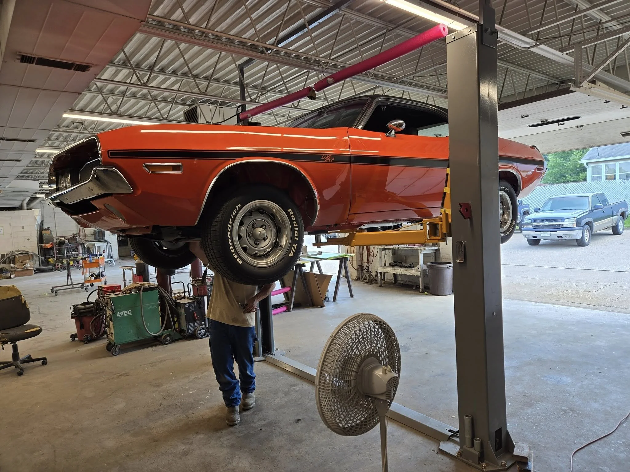 A red vintage car is lifted on a hydraulic lift inside a garage or auto repair shop. A person is working underneath the car. There is a large fan and various tools and equipment in the garage.