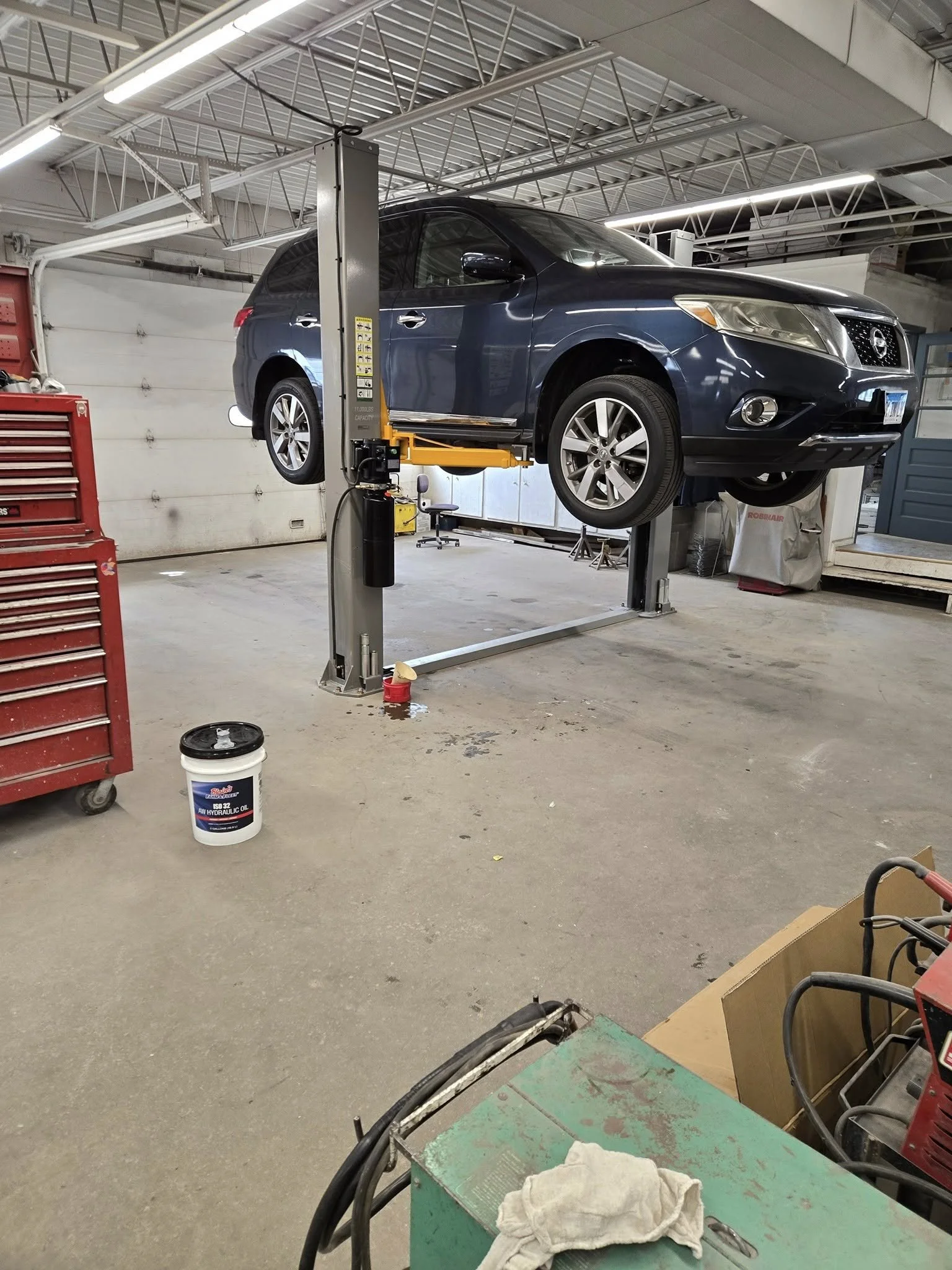 A black SUV is elevated on a hydraulic lift inside an automotive repair shop, with tools and equipment surrounding the area.