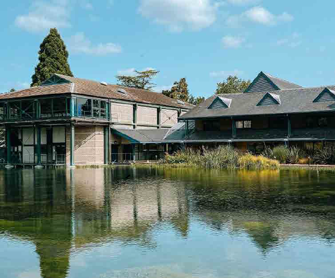 Large modern house with multiple levels and a unique roof design, situated next to a body of water, with trees and blue sky in the background.