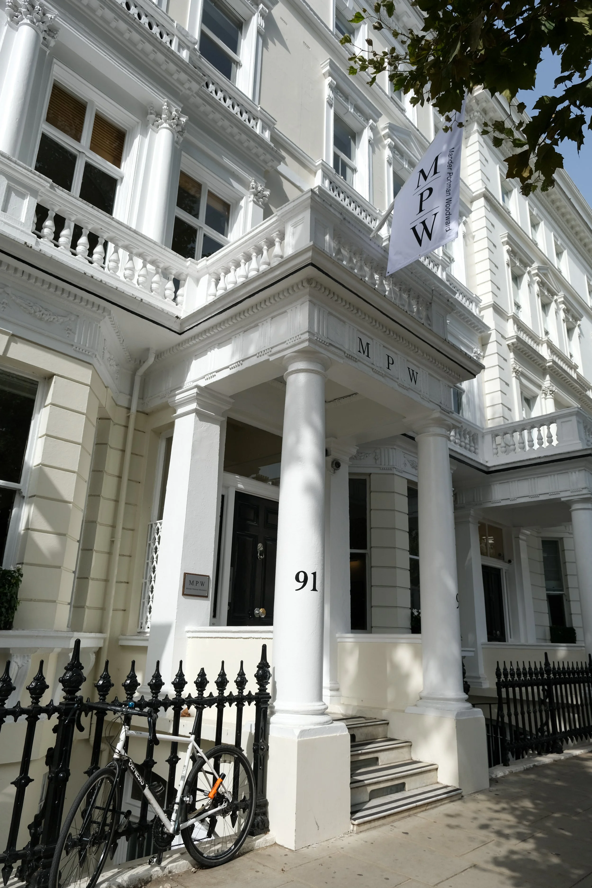 White multi-story building with classical architectural details, columns, balustrades, and decorative molding. A flag with 'MPW' and 'Wander Pohlman Woodward' hangs from the upper balcony. The building's street number is 91, and a black bicycle is parked against the fence in front.