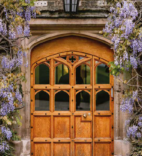 A wooden arched door set in stone, surrounded by purple flowering plants.
