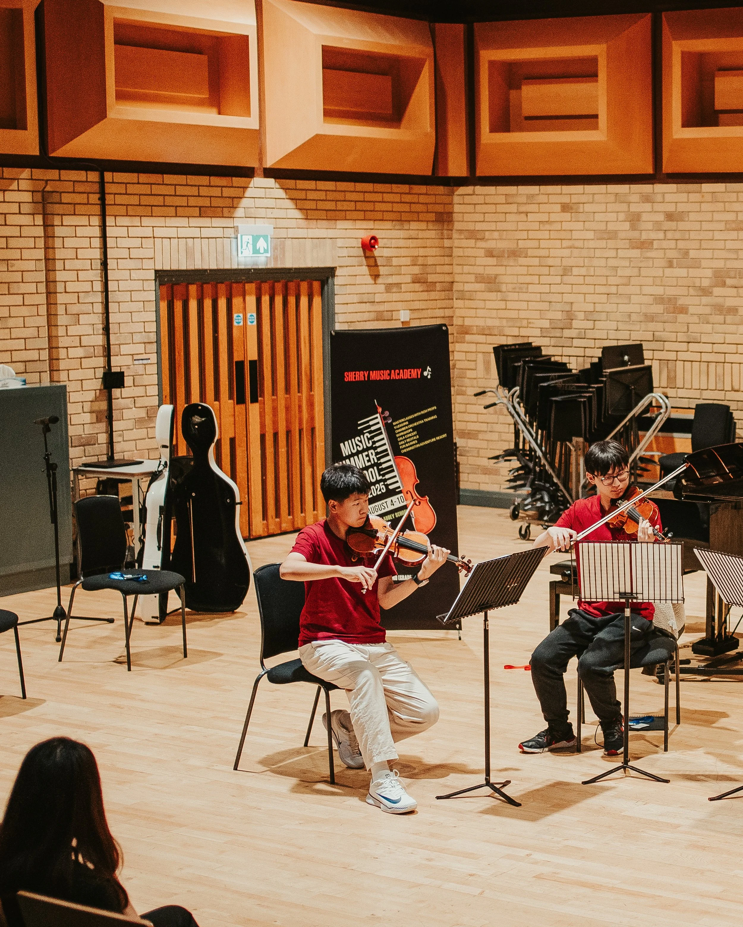 Two young musicians, one male and one female, playing violins during a performance on a stage with a brick wall background. There are music stands and chairs around them, and a sign indicating an emergency exit is visible above a door behind them.