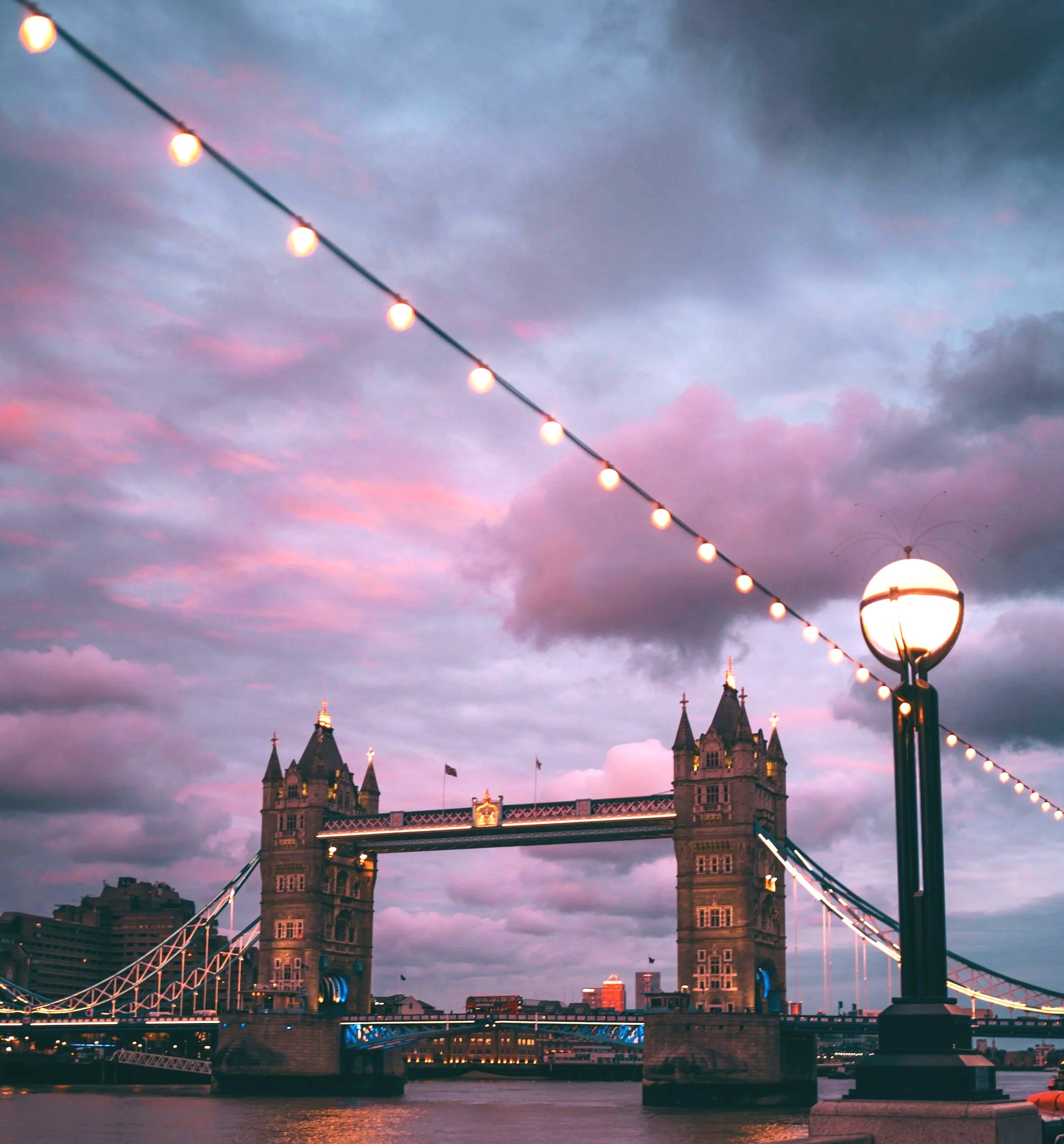 Sunset view of Tower Bridge in London with cloudy sky and string lights in the foreground.