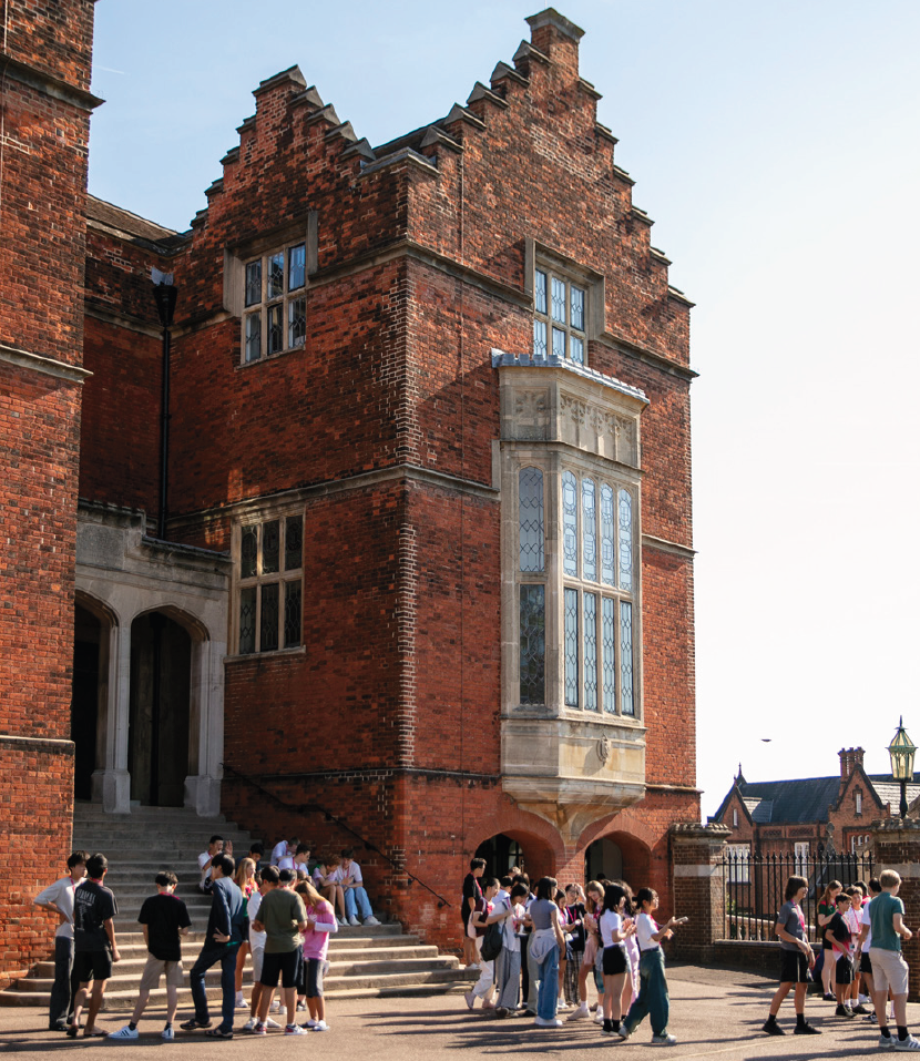 School children gather outside a historic brick building with a large bay window and stone archway.