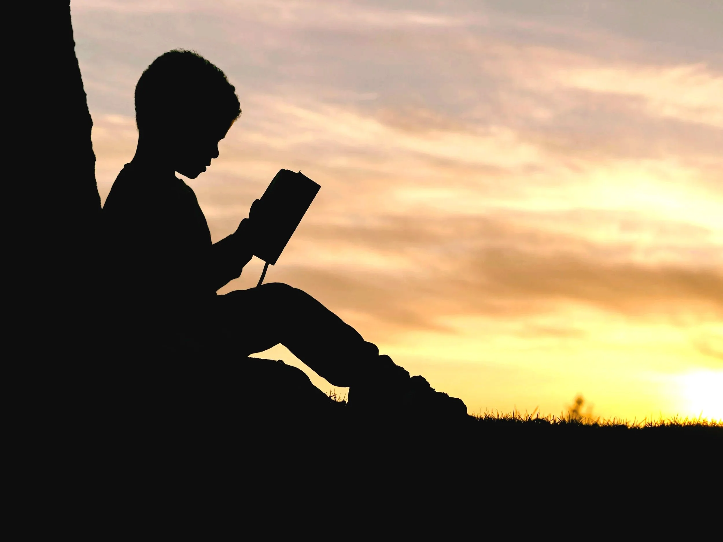 Silhouette of a child reading a book while sitting against a rock on a hillside during sunset.