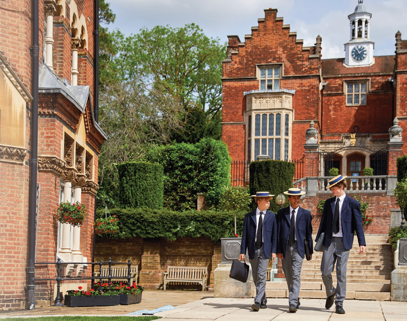 Three young men in suits and straw boater hats walking on a stone pathway in front of a historic red brick building with green trees, potted plants, and benches.