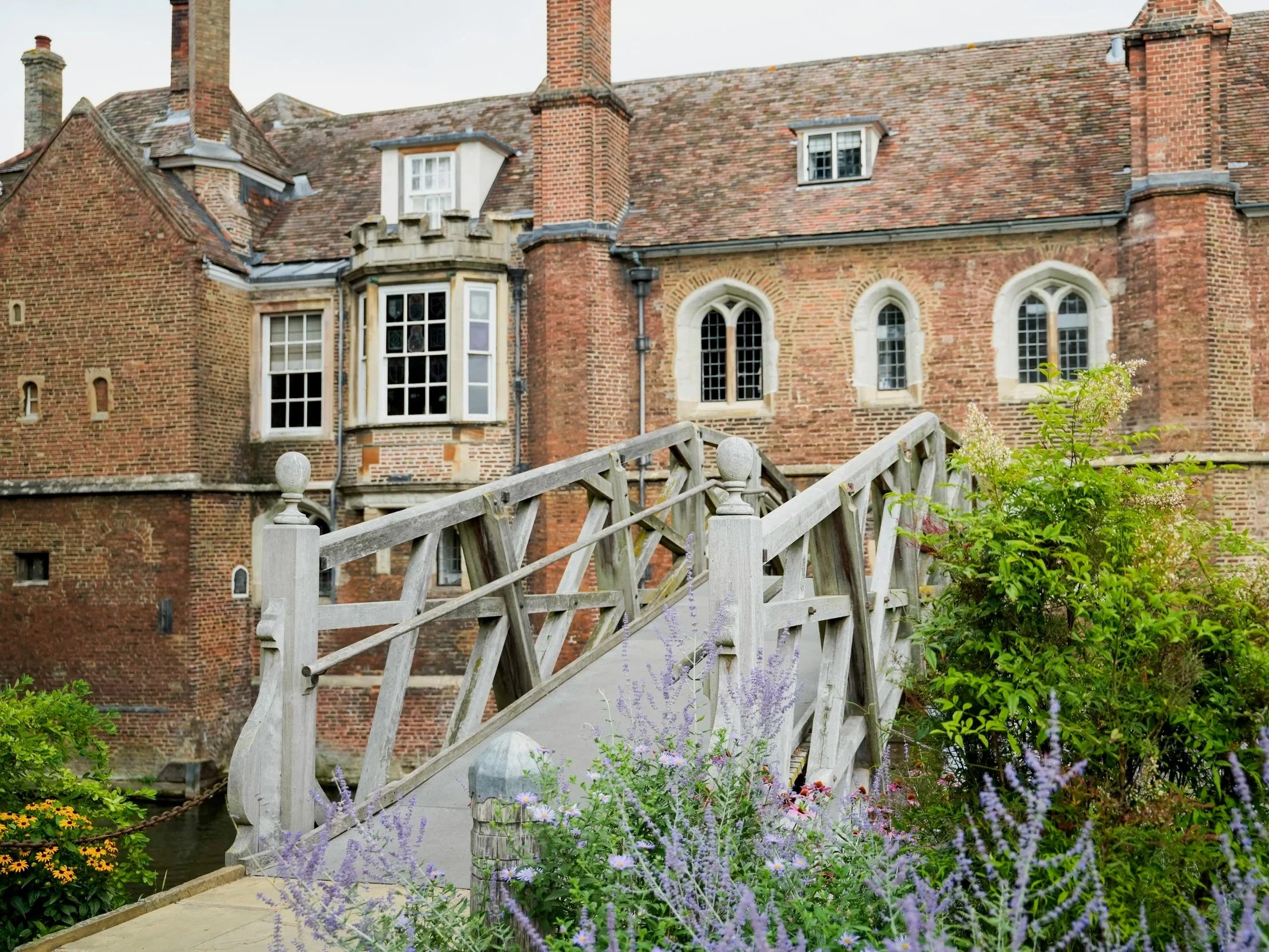 A brick house with multiple chimneys, windows, and a small castle-like turret, seen from a garden with a wooden footbridge crossing over a pond or stream and surrounded by blooming flowers and green foliage.