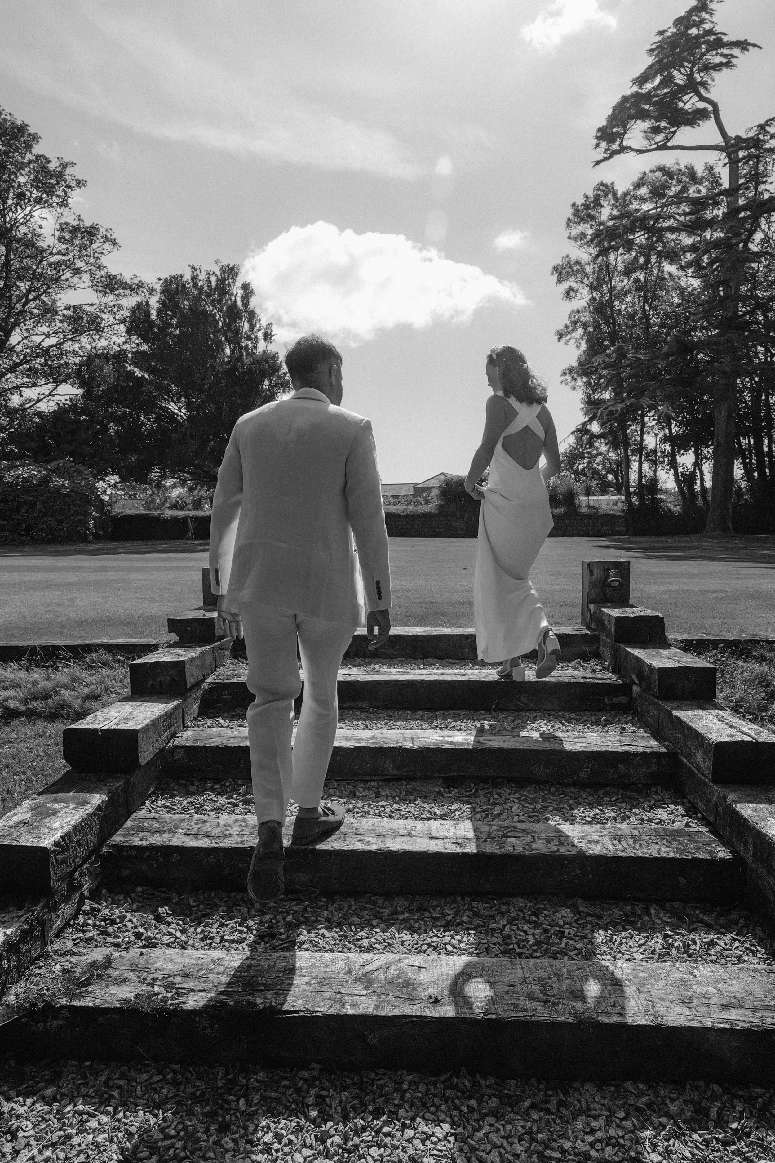 A black and white photo of a man and woman walking up wooden stairs in an outdoor park or garden with trees and a partly cloudy sky in the background.