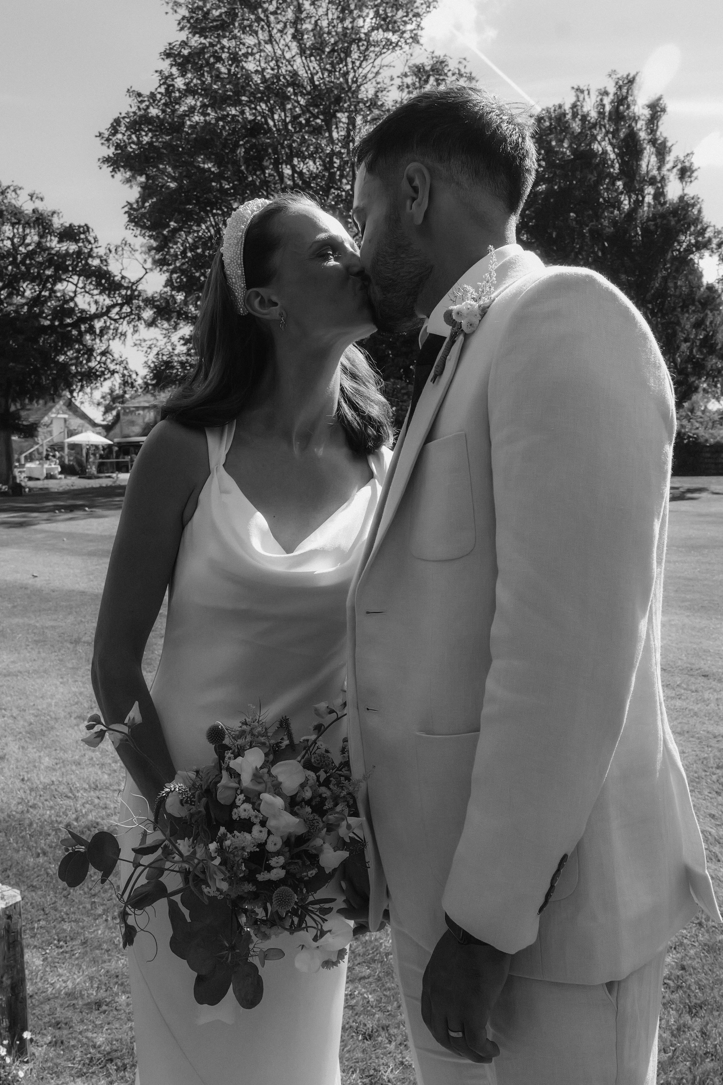 A black and white photo of a bride and groom sharing a kiss outdoors, with trees and a small building in the background. The bride is holding a bouquet of flowers, and the groom is dressed in a light-colored suit with a boutonniere.