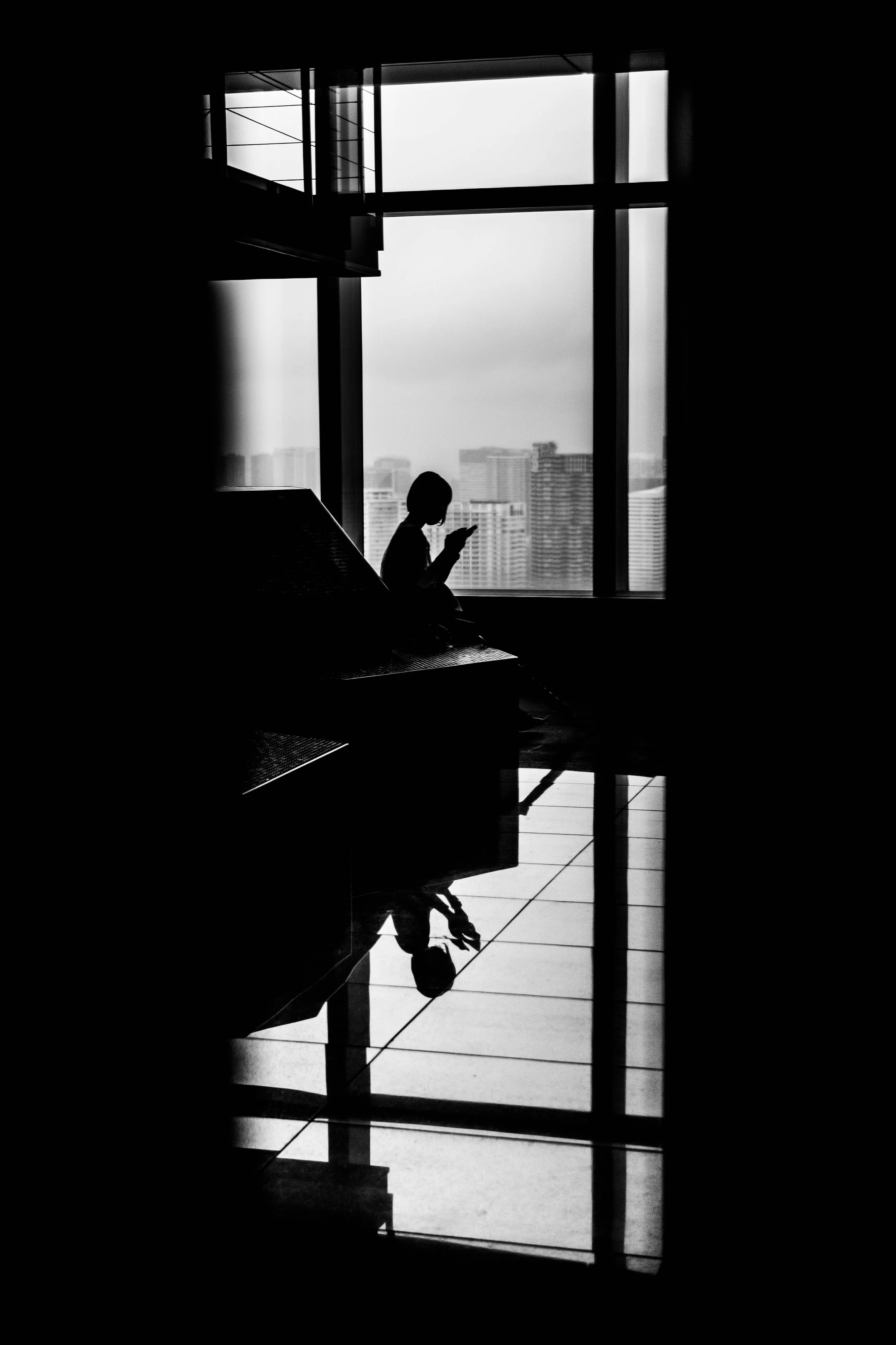 Silhouette einer Person, die auf einer Bank in einem modernen Gebäude sitzt und ein Smartphone benutzt, mit Blick auf eine Stadt im Hintergrund, reflektiert auf dem Boden, schwarz-weiß