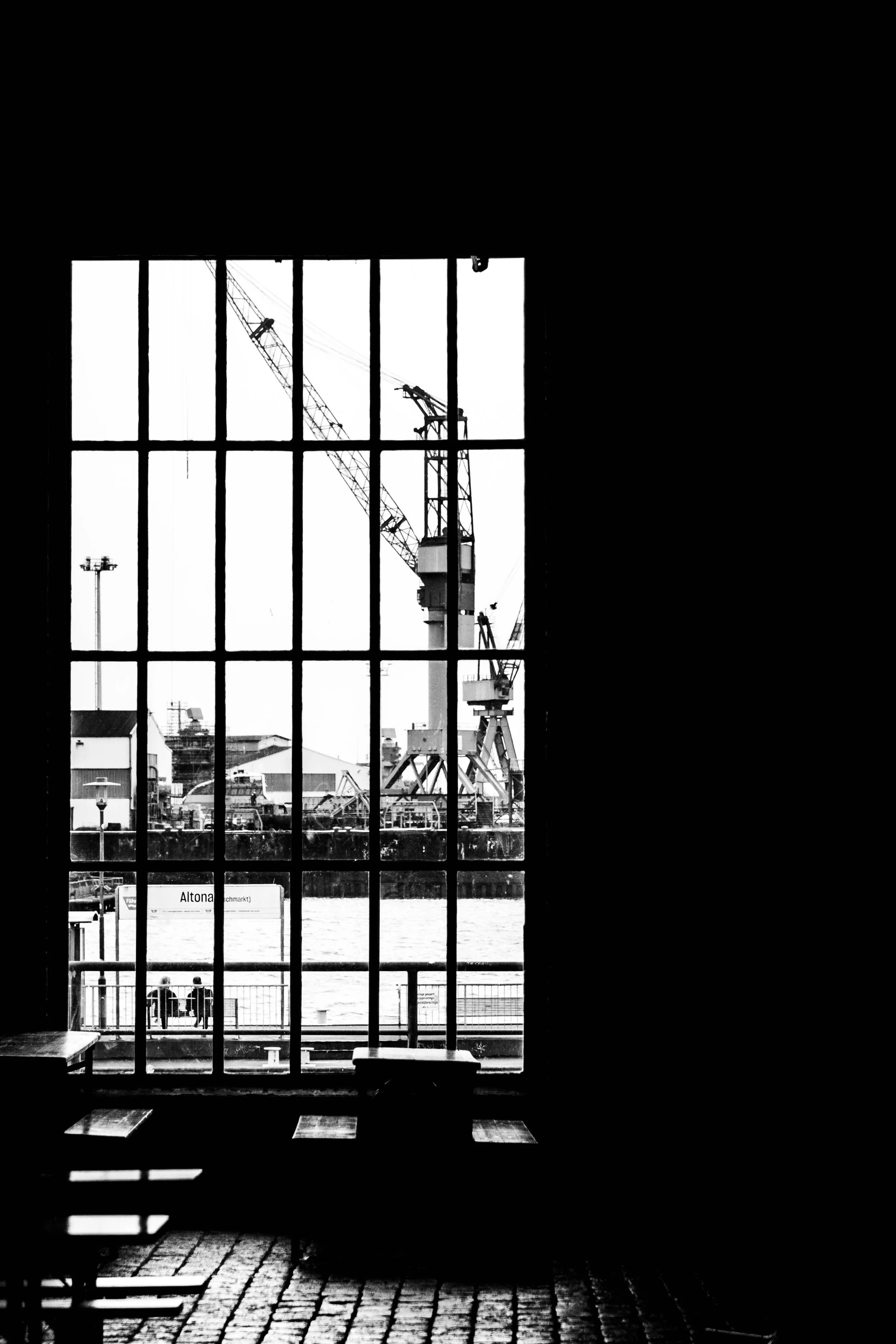 Blick durch ein Gitterfenster auf einen Hafen mit Kränen und Wasser im Hintergrund, schwarz-weiß