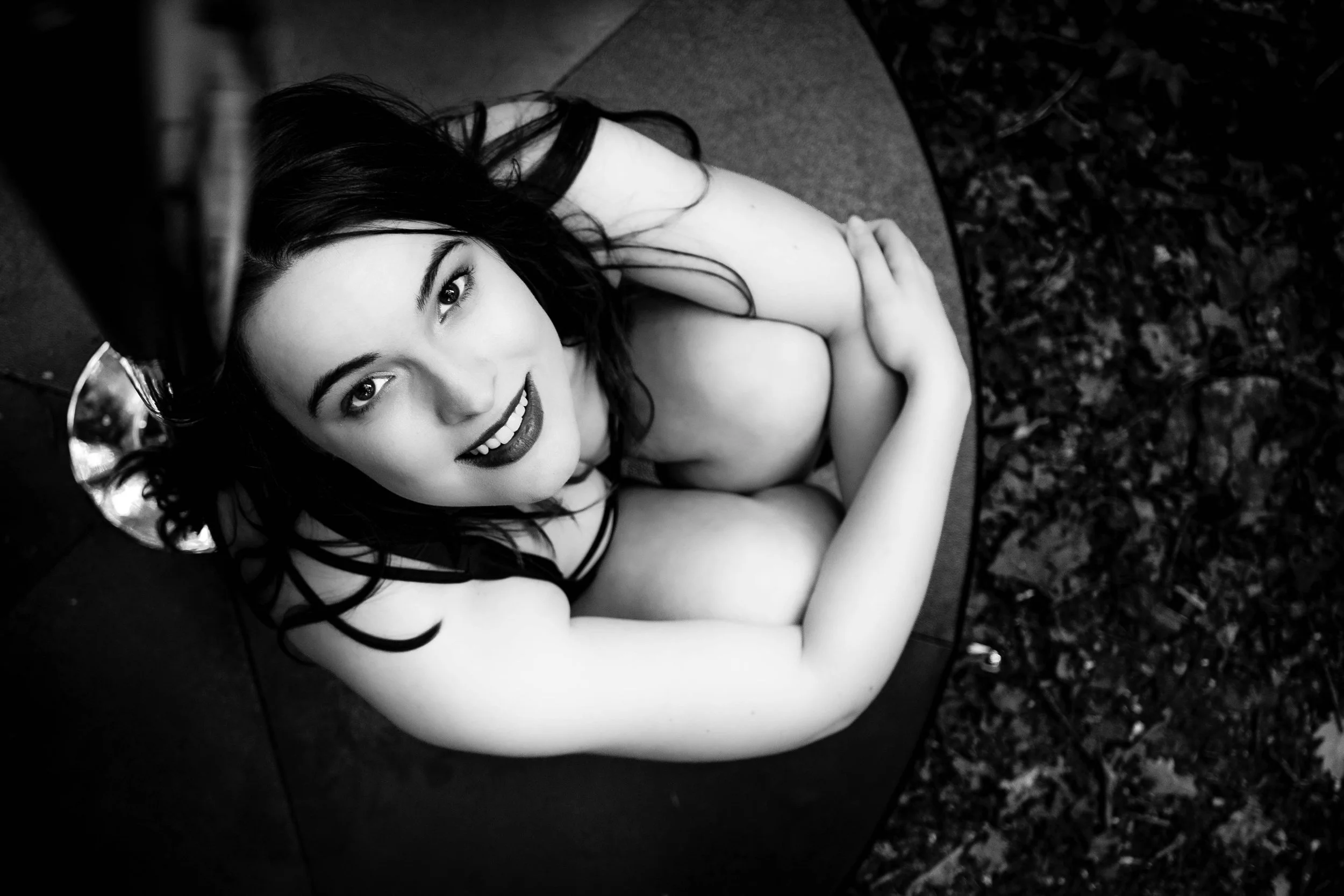 Schwarzweiß-Fotografie einer lächelnden Frau, die auf einem runden, sandfarbenen Sitz im Freien sitzt, mit dunklem Haar, Kleiderträger sichtbar, umgeben von Natur mit Blättern auf der Erde.