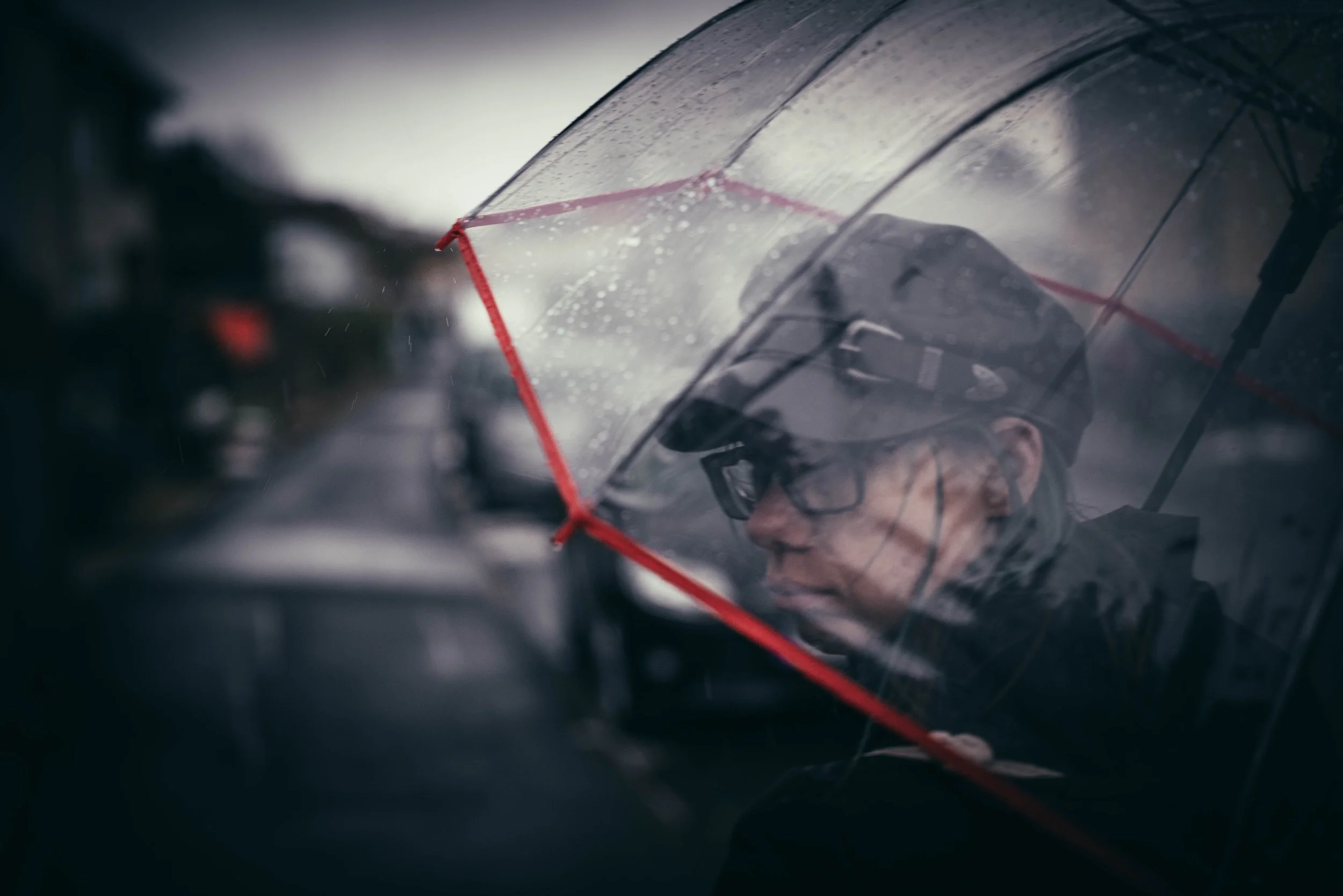Ein Mensch mit Regenjacke, Mütze und Brille hält einen transparenten Regenschirm bei Regen.
