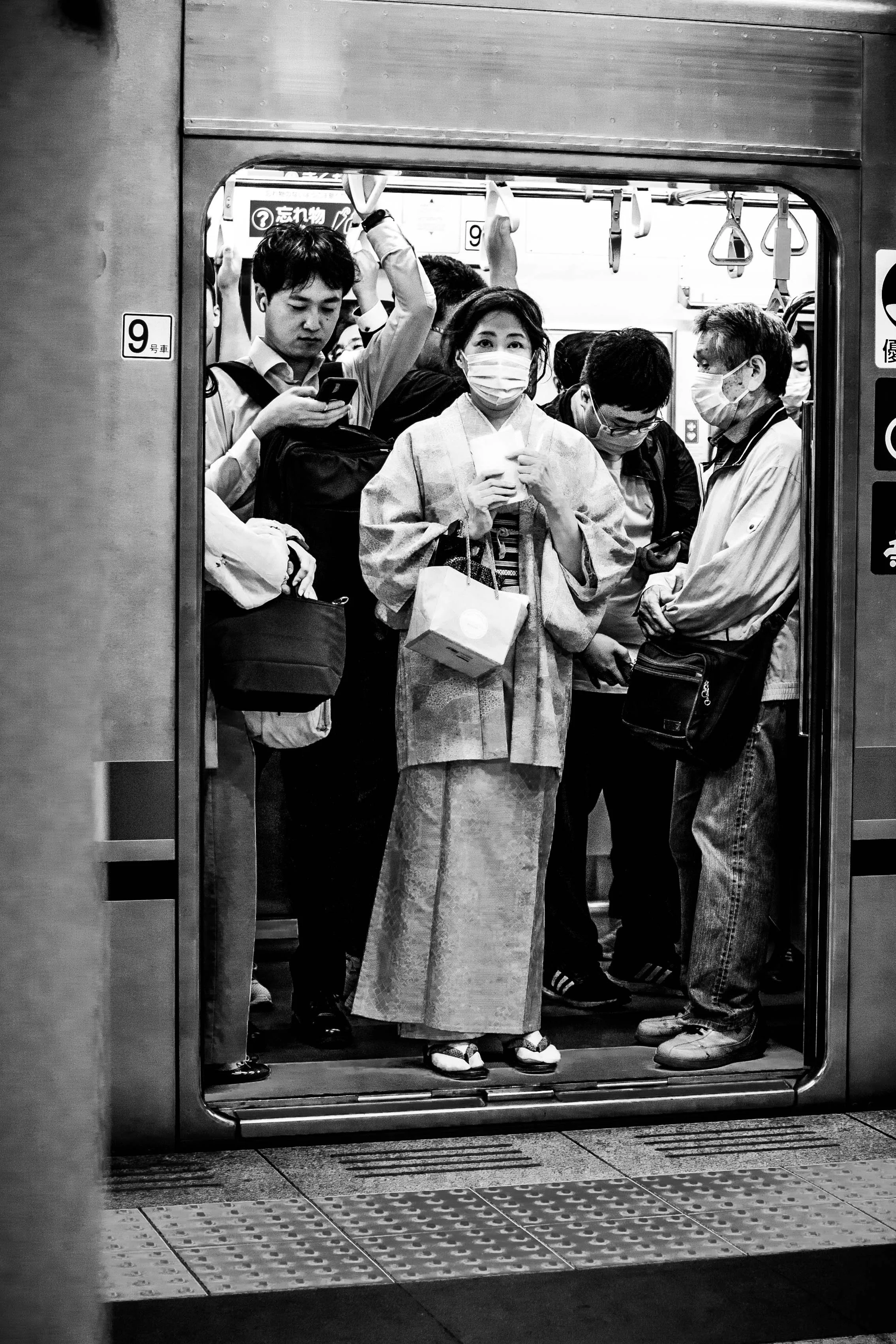 Menschen stehen in einer U-Bahn, einige tragen Masken, eine Frau in einem Kimono hält einen Becher, alle schauen nach unten oder auf ihre Telefone.