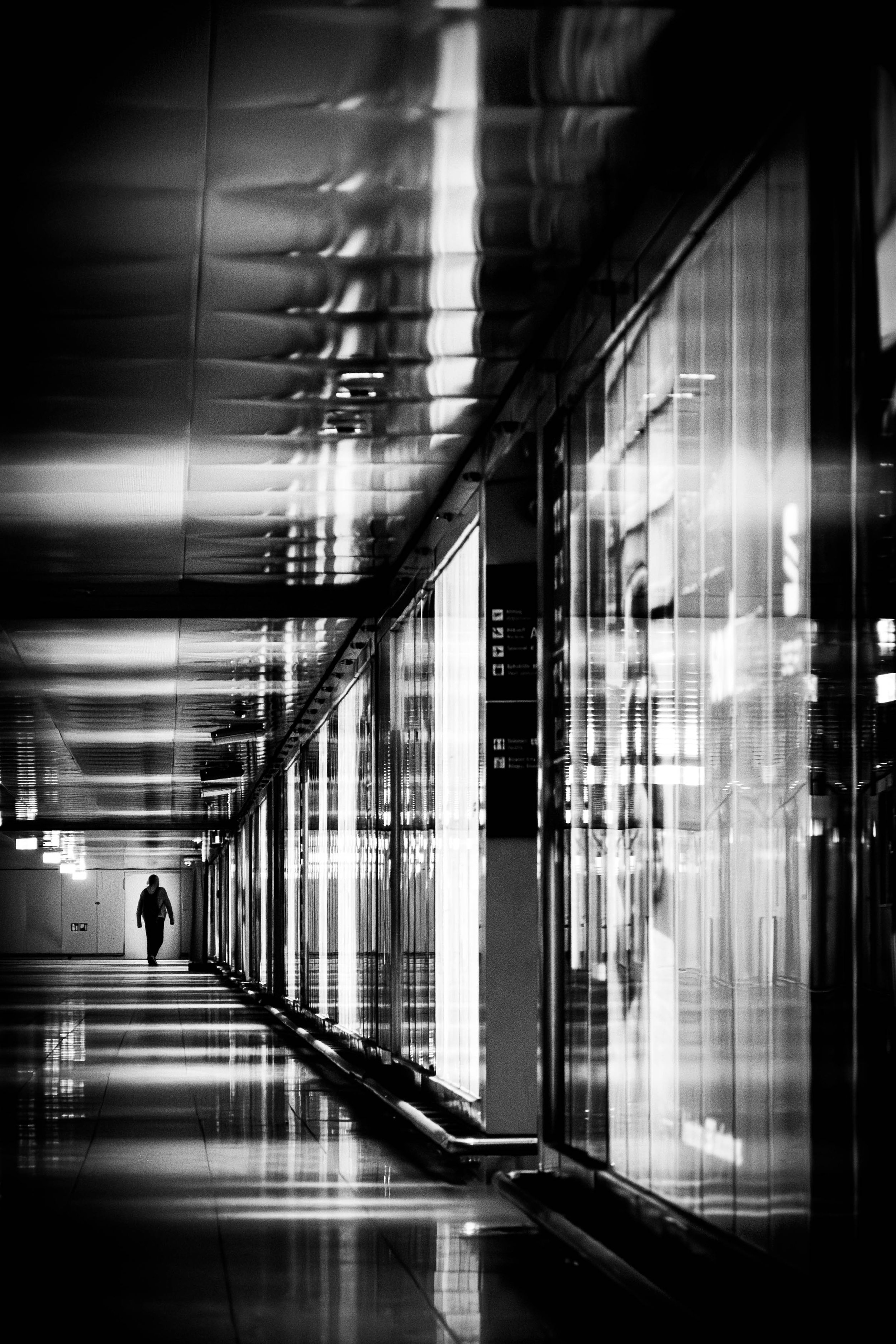 Ein einsamer Mensch geht durch einen dunklen, reflektierenden Korridor mit metallischen Wandverkleidungen im Schwarz-Weiß-Foto.