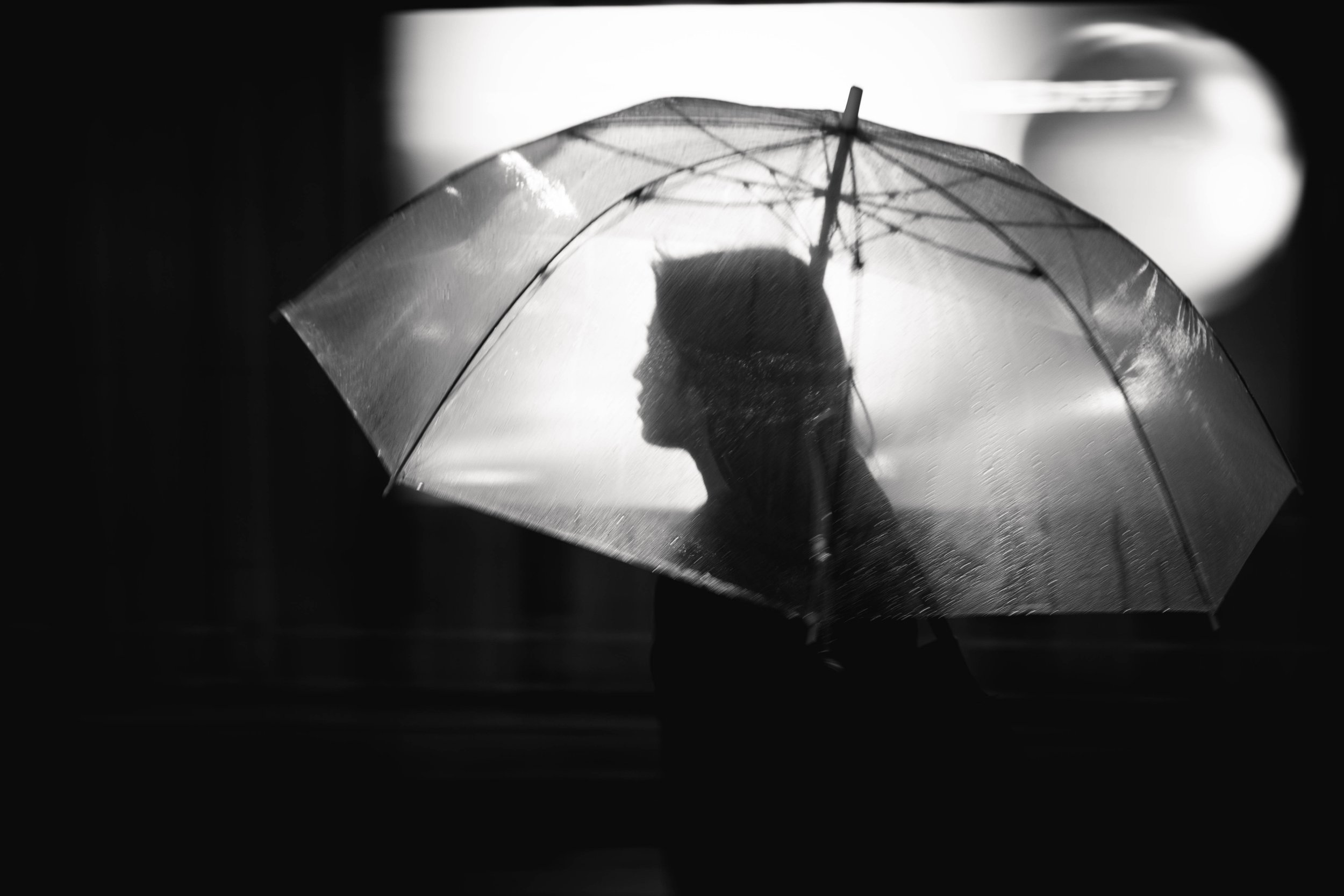 Schwarz-weiß Foto einer Person, die einen durchsichtigen Schirm hält, während es regnet. Die Person ist seitlich im Fokus, während im Hintergrund verschwommenes Licht zu sehen ist.