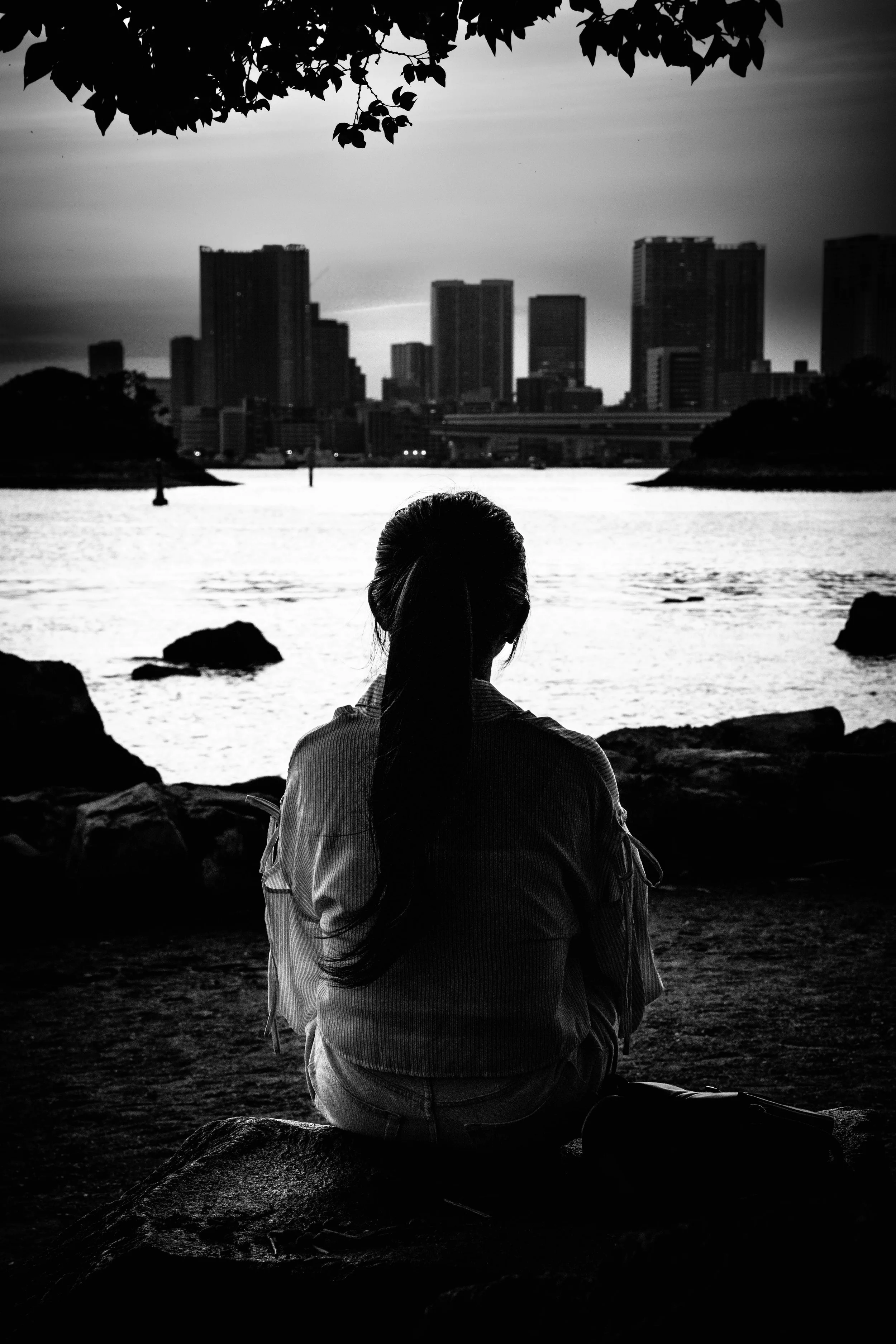 Eine Person sitzt am Wasser und blickt auf eine Stadt im Sonnenuntergang, alles in Schwarz-Weiß.