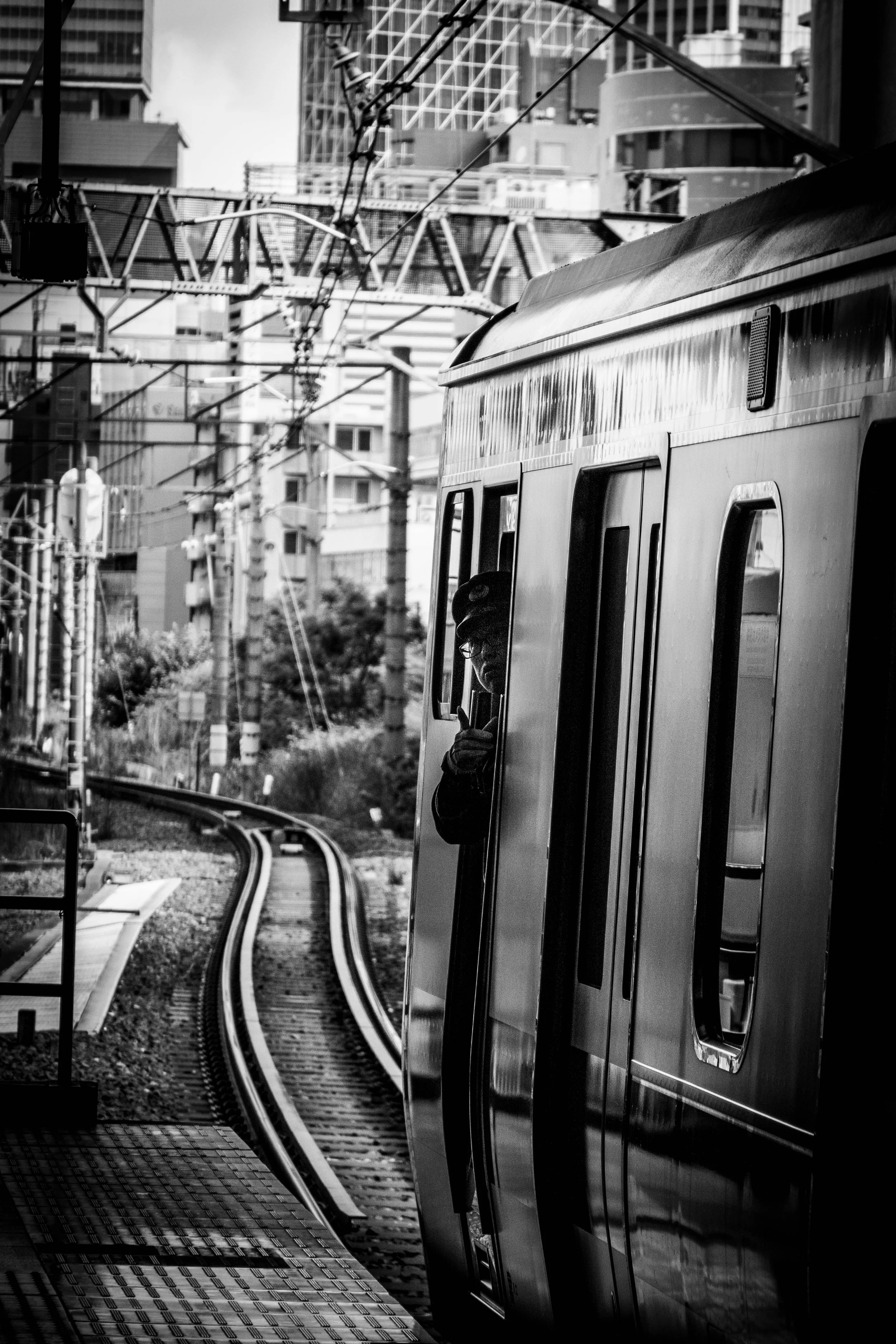 Ein Mann schaut aus einem U-Bahnhof-Fenster heraus, während ein Zug vorbeifährt, umgeben von einer städtischen Kulisse mit Hochhäusern und elektrischen Leitungen in Schwarz-Weiß.