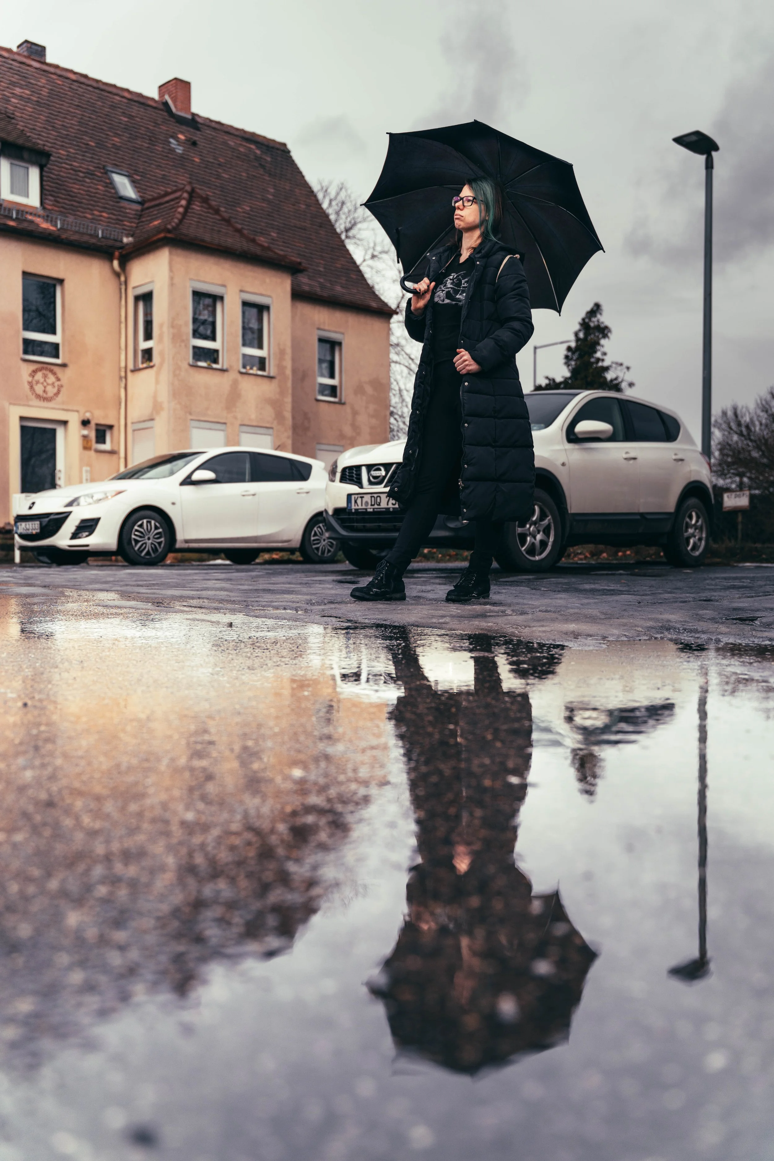 Frau mit Regenschirm geht auf regennasser Straße mit Pfützen, Häuser und geparkte Autos im Hintergrund, bewölkter Himmel.