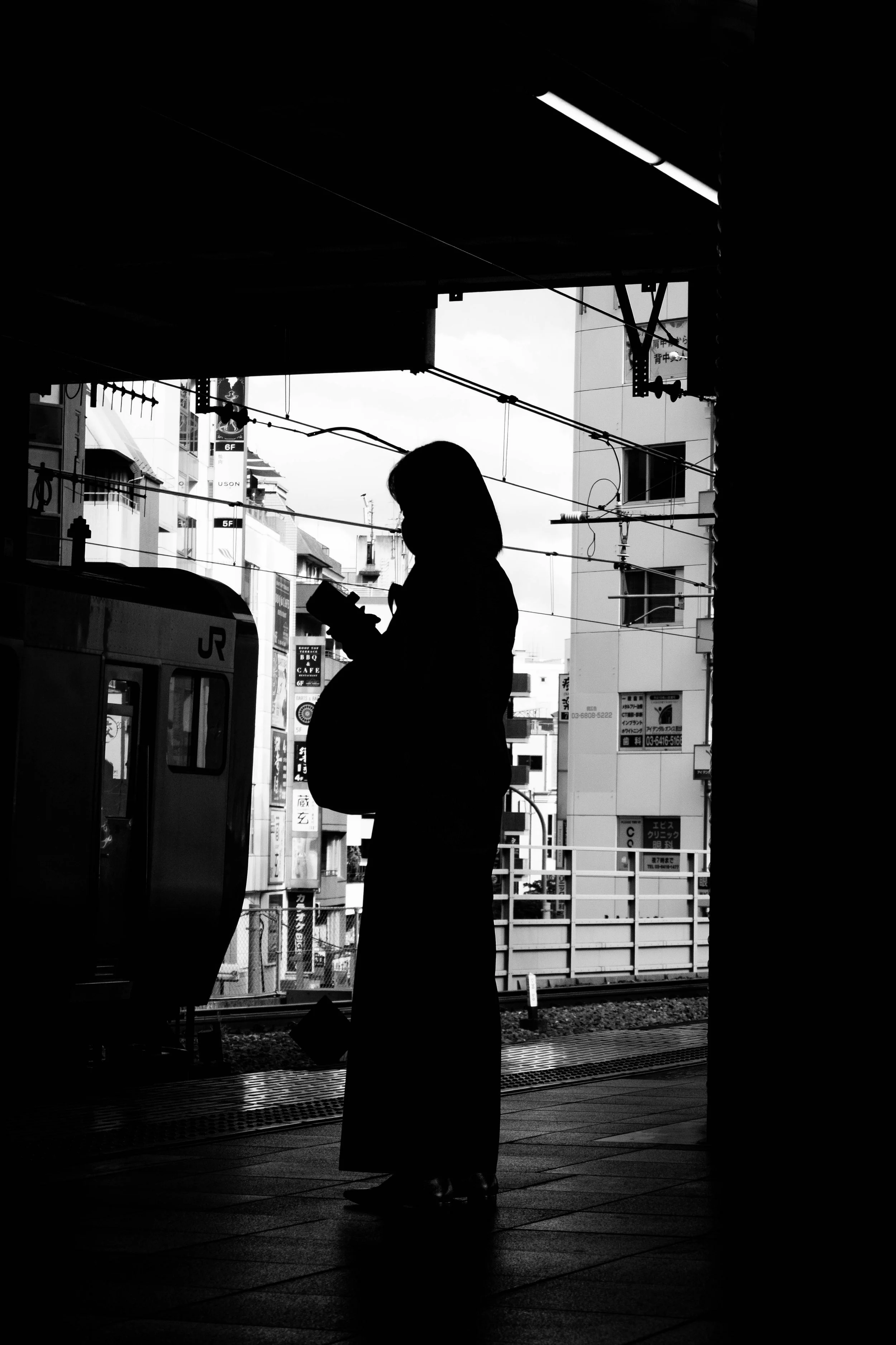 Silhouette einer schwangeren Frau, die auf einem Bahnsteig steht und ein Smartphone benutzt, vor dem Hintergrund eines städtischen Bahnareals in Schwarz-Weiß.
