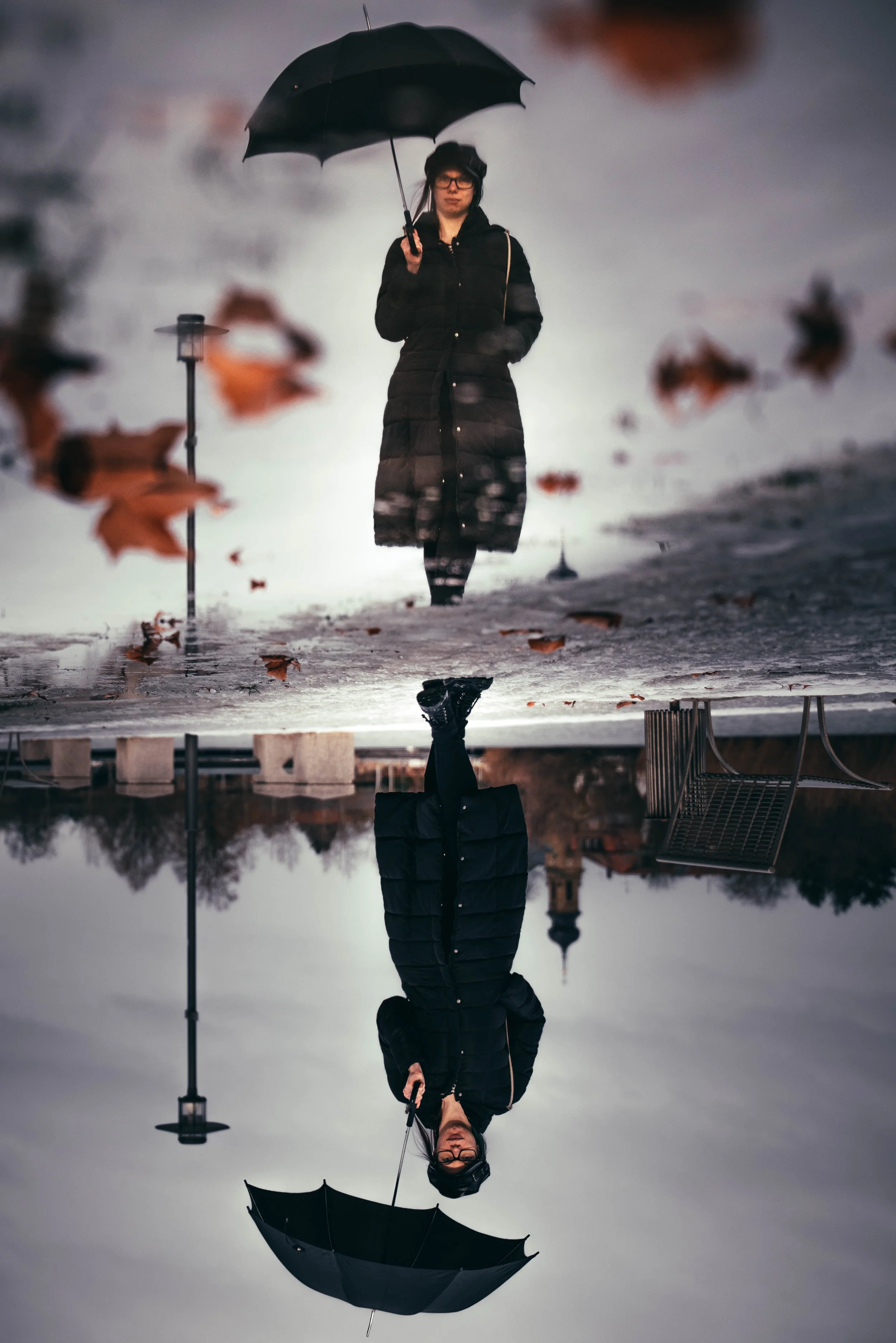 Eine Frau in einem schwarzen Mantel und mit Regenschirm steht auf einer spiegelnden Wasserfläche, die den Himmel und die Umgebung widerspiegelt, mit Blättern im Wasser.