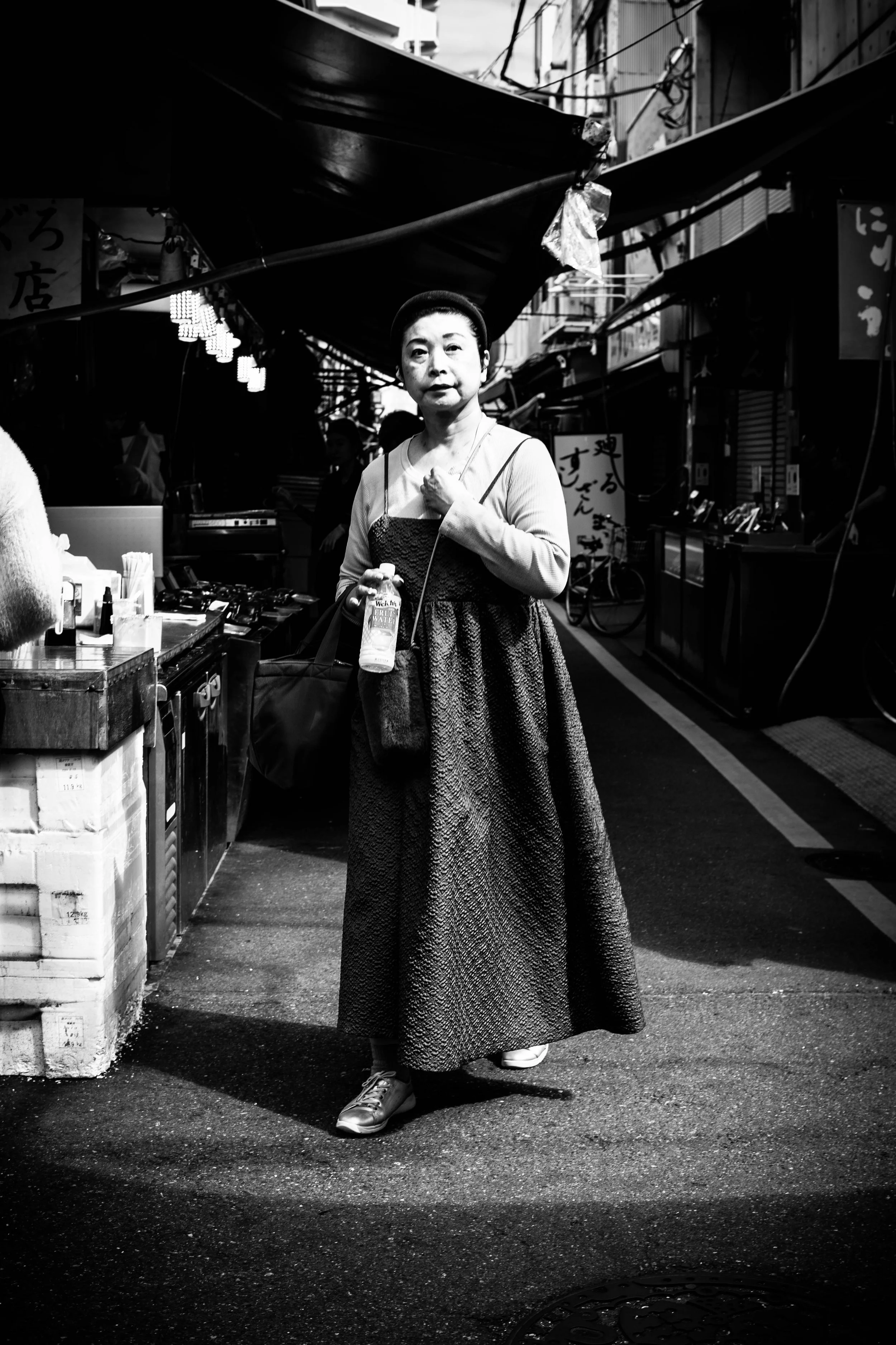 Eine asiatische Frau trägt ein langes Kleid und hält eine Wasserflasche, während sie durch einen belebten Markt in Tokyo geht.