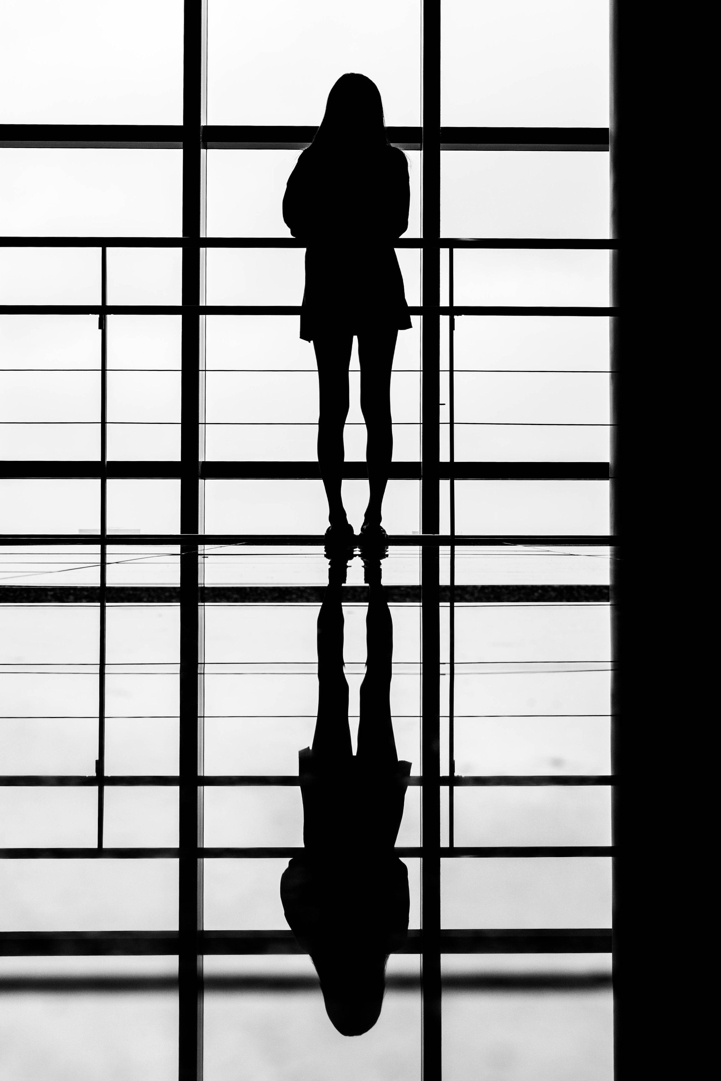 Silhouette einer stehenden Frau vor großen fenstern, deren Reflexion auf dem Boden sichtbar ist.