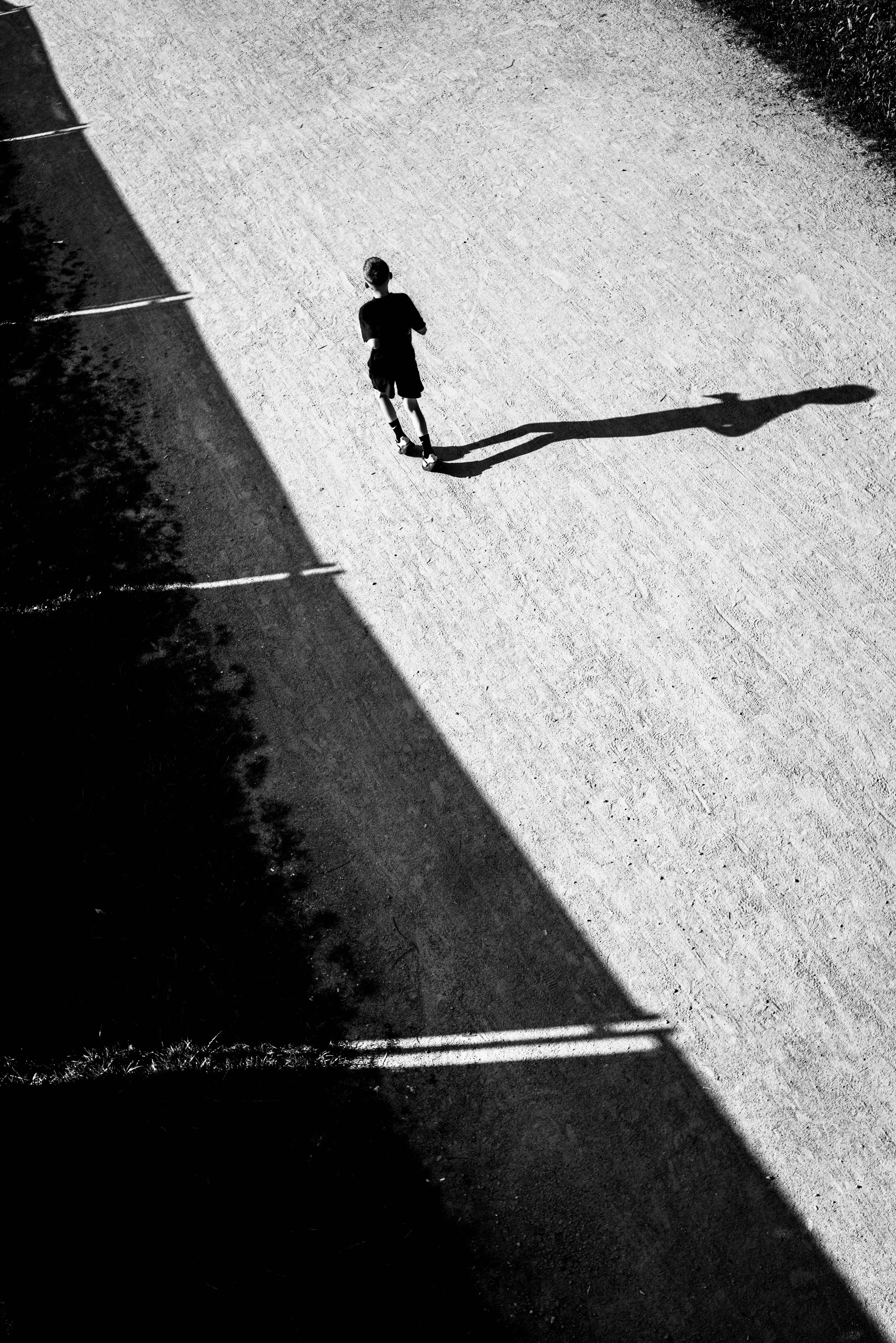 Ein Junge läuft auf einer hellen, schotterbedeckten Oberfläche, während sein langer Schatten von der Sonne auf den Boden geworfen wird. Ein dunkler Schattenbereich befindet sich auf der linken Seite des Bildes.