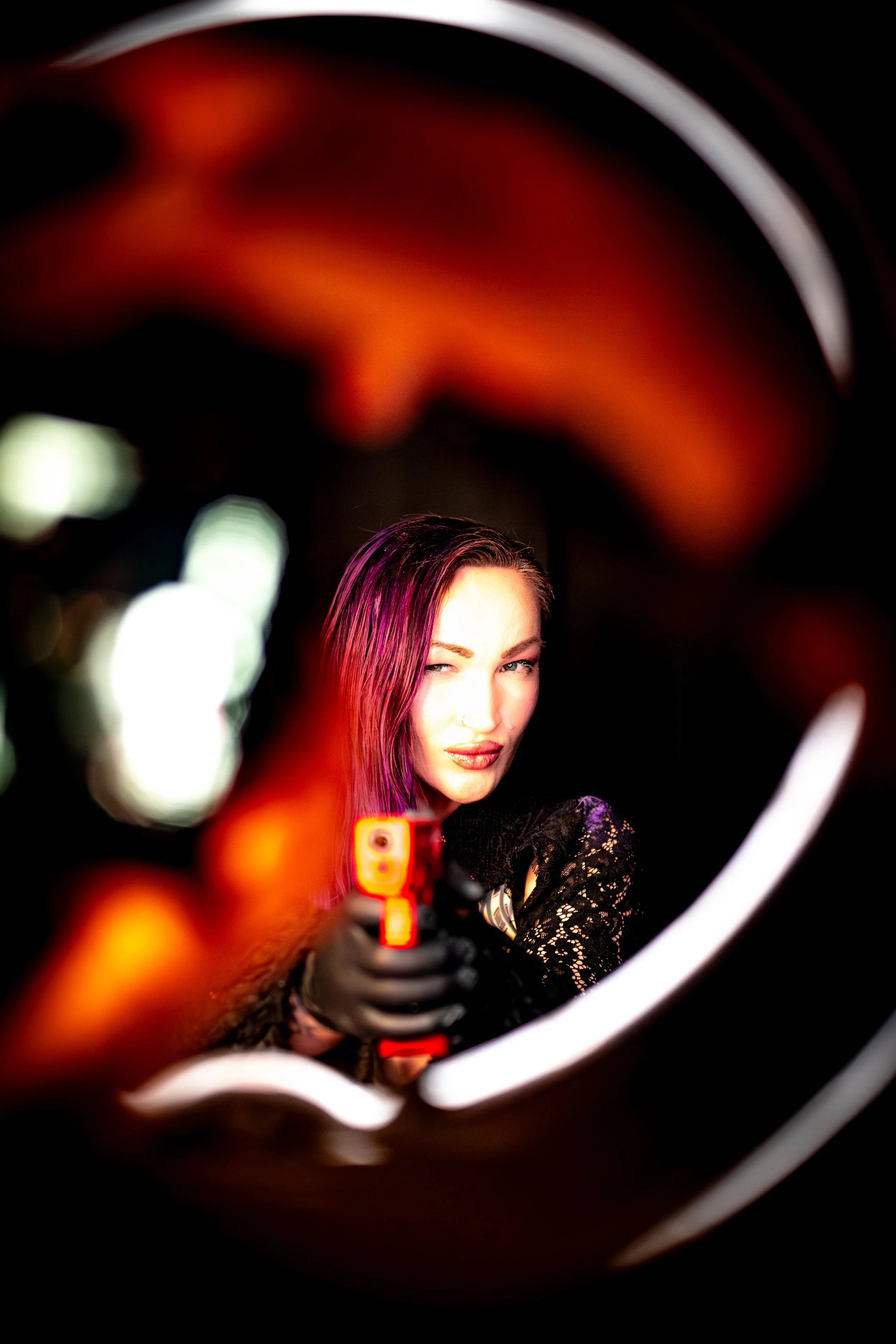 Frau mit lila Haaren, die in die Kamera hält eine Spielzeugwaffe, umgeben von schwarzen und roten Elementen.
