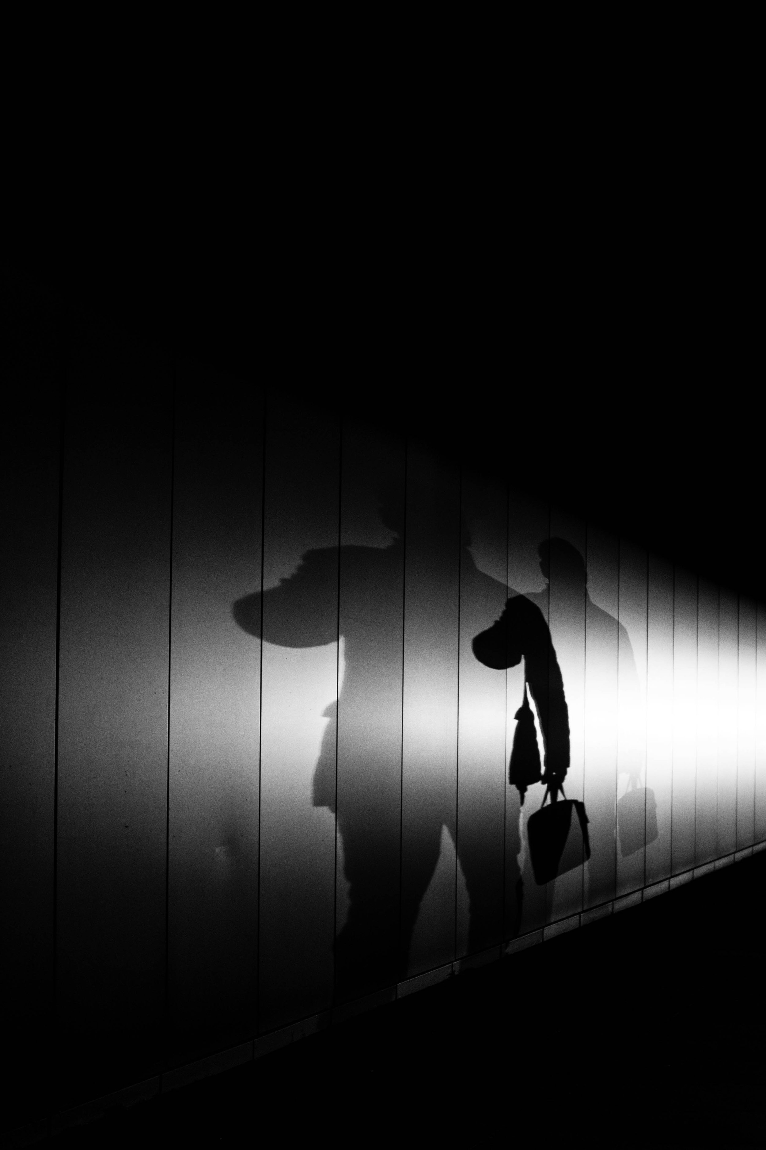 Schwarze Silhouetten zweier Menschen mit Koffern und Taschen, die Schatten an einer beleuchteten Wand hinterlassen, in schwarz-weiß.