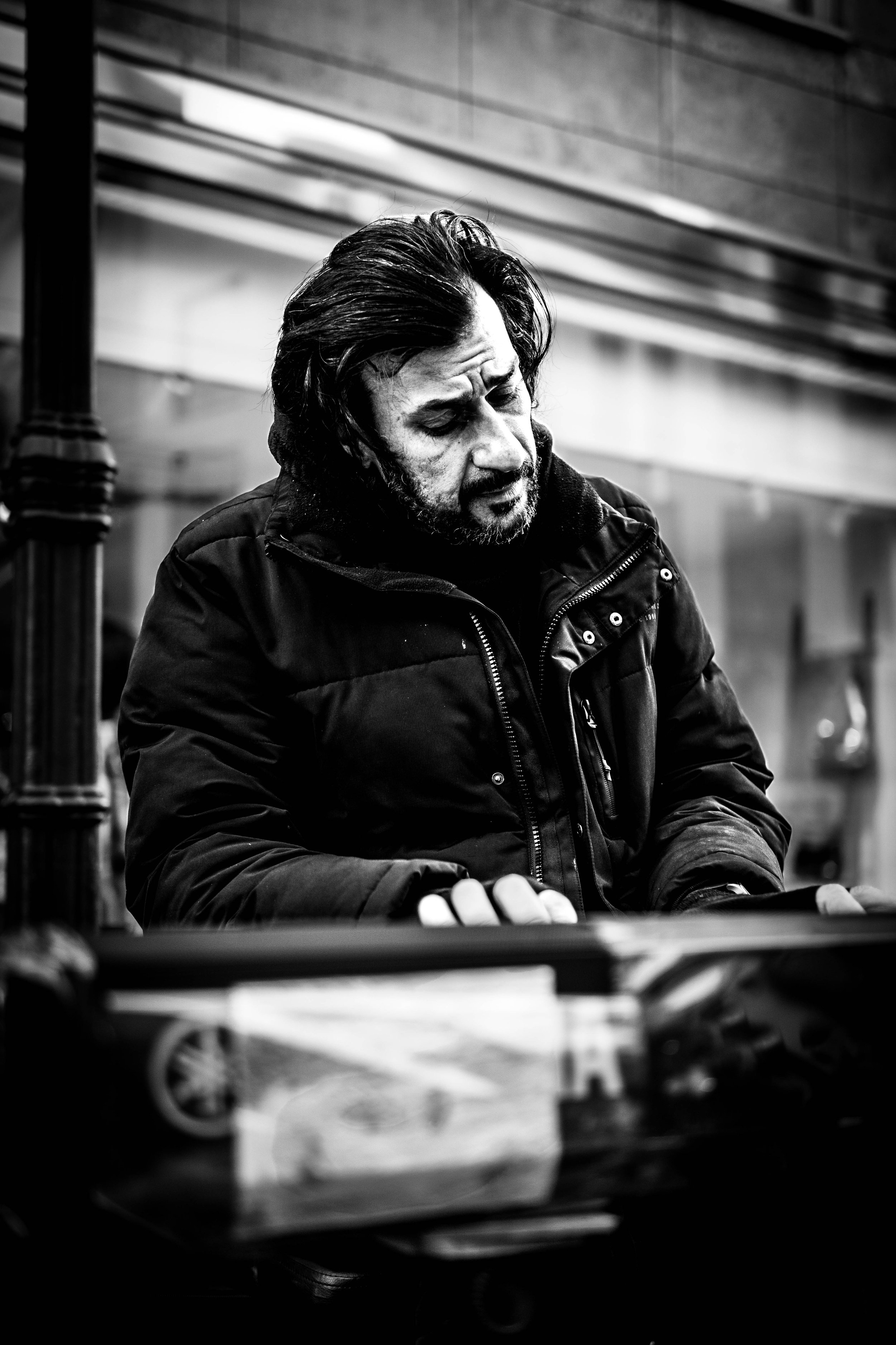 Schwarz-Weiß-Foto eines Mannes mit dunklen, langen Haaren und Gesichtsbart, der nachdenklich schaut, während er an einem Gegenstand arbeitet, in einer urbanen Umgebung.