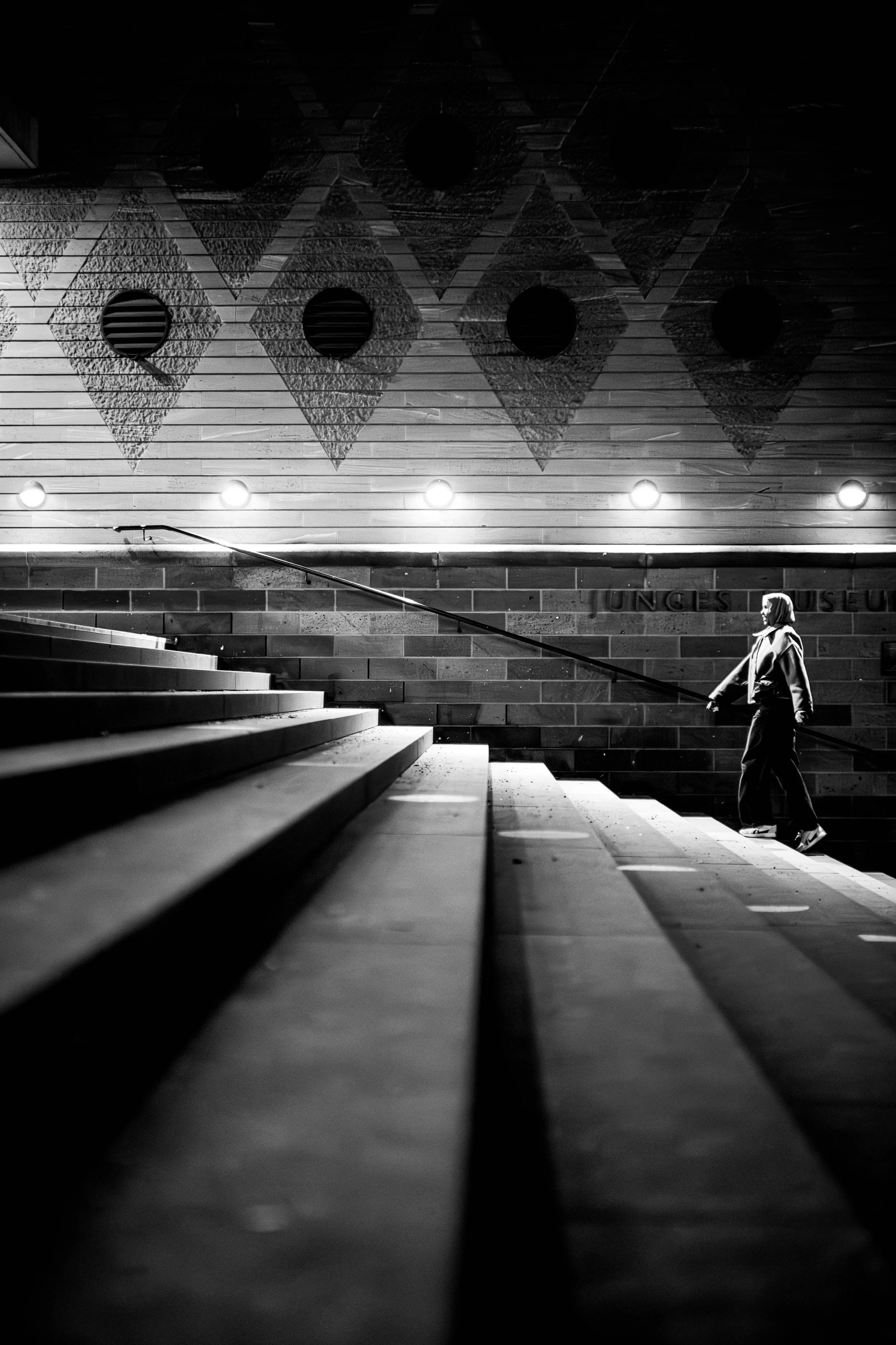 Ein schwarz-weiß Foto eines Mannes, der eine Treppe hinaufgeht, im Hintergrund eine Wand mit geometrischen Mustern und Beleuchtung.