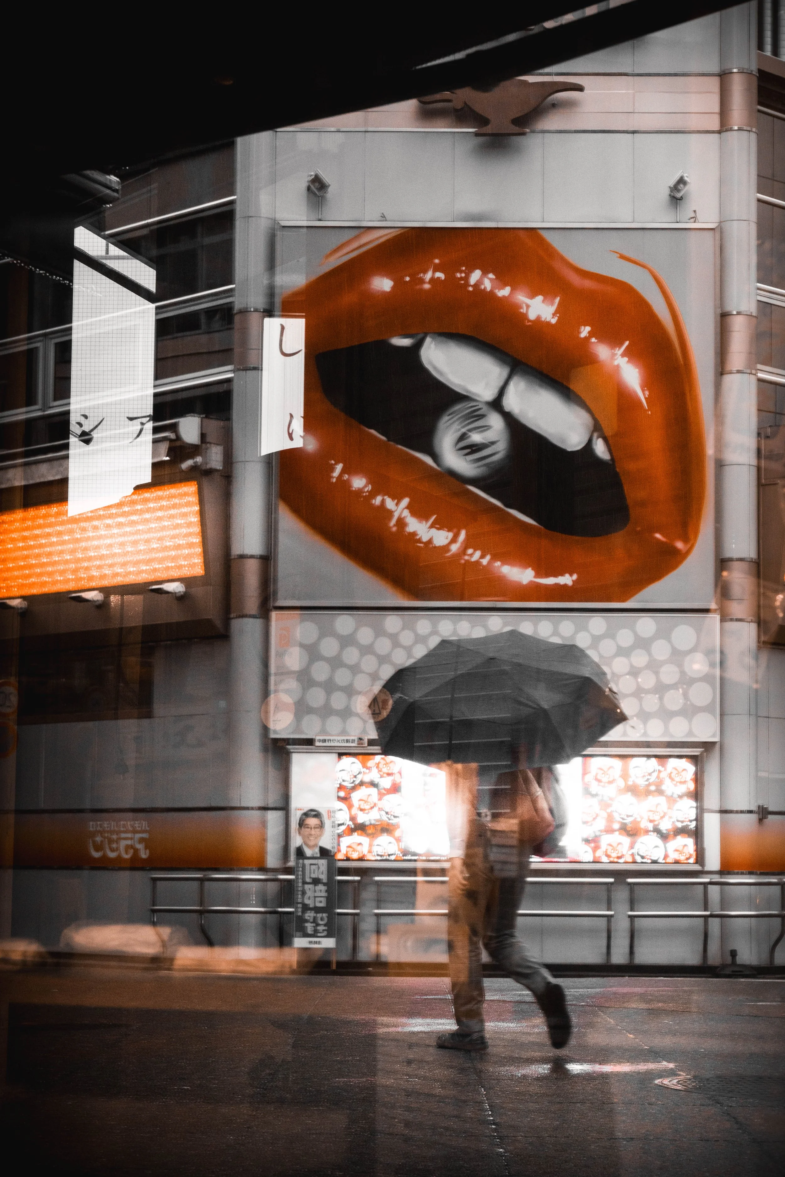 Eine Person mit Regenschirm geht an einem Gebäude vorbei, das mit großen Werbeplakaten mit einem roten Lippen-Motiv und einem Damenporträt bedeckt ist, bei Regen in einer Stadt.