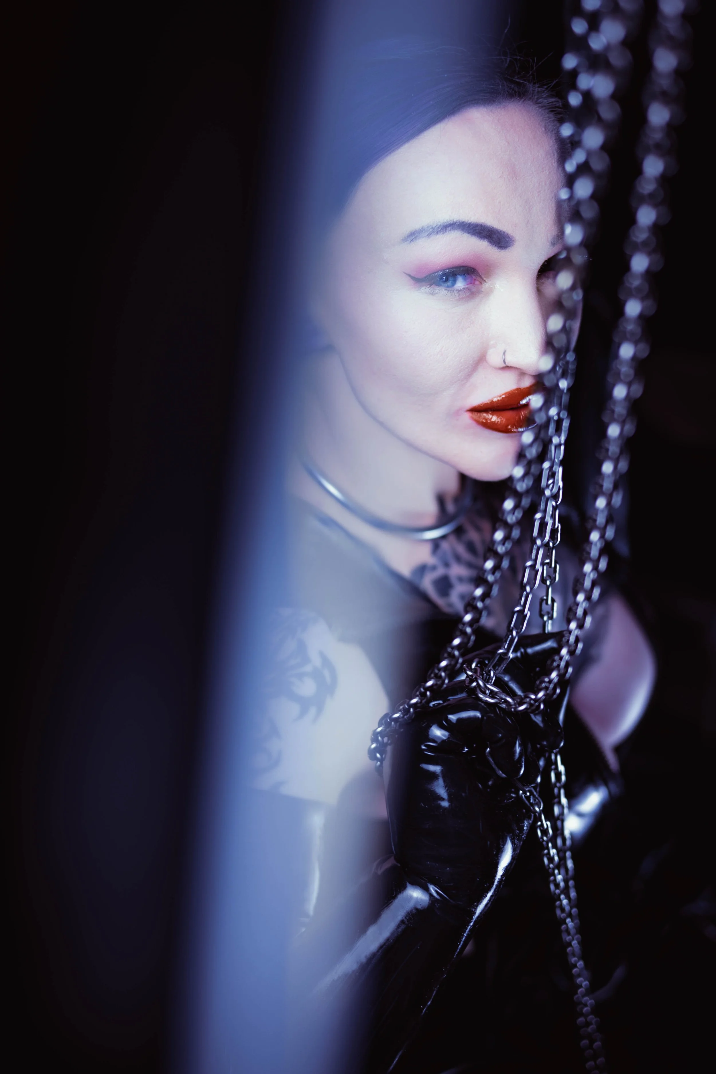 Nahaufnahme eines Models mit dunklem Haar, rotem Lippenstift, Ketten und Lederhandschuhen, das durch einen schmalen Spalt zwischen dunklen Vorhängen betrachtet wird.