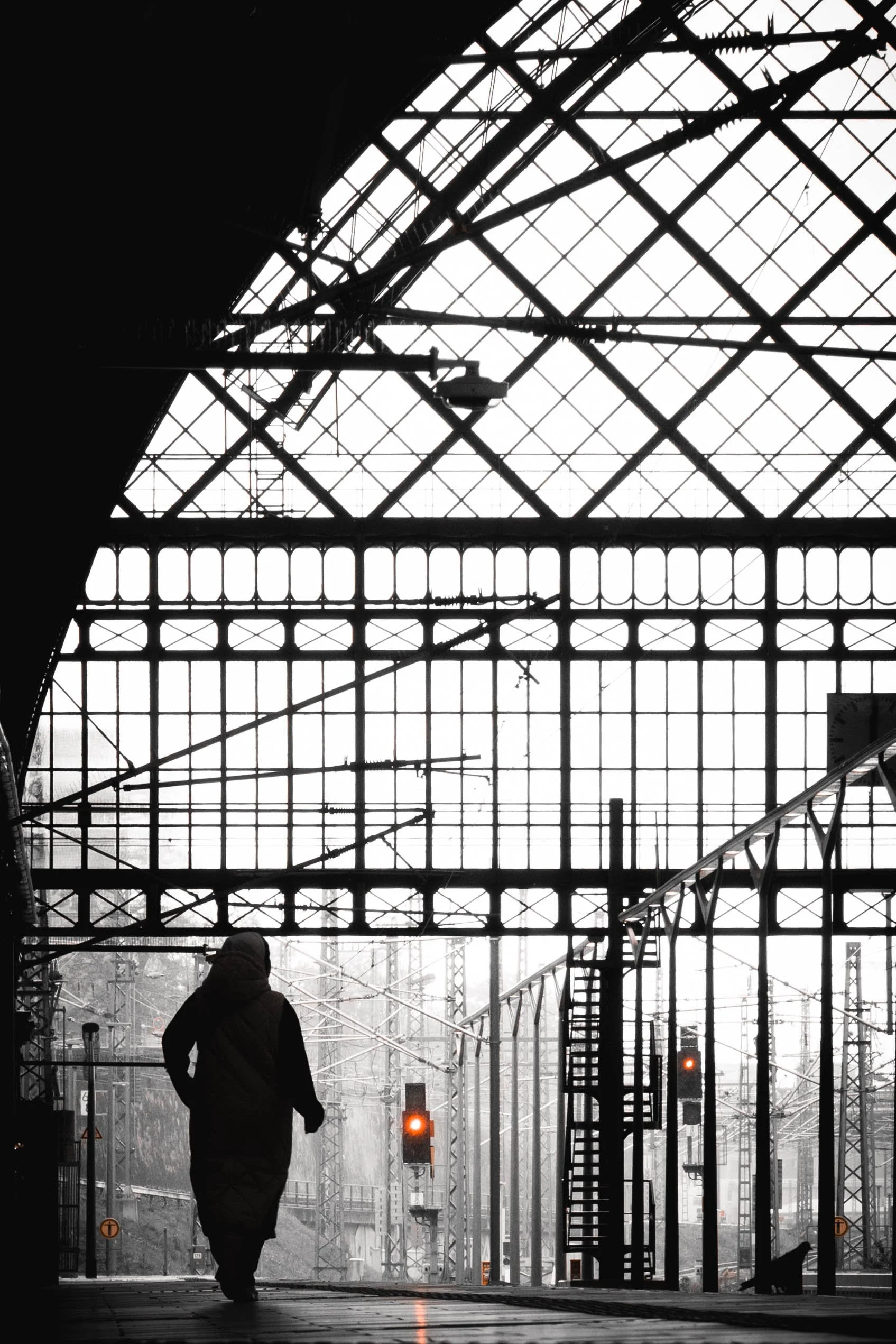 Ein Mensch läuft in einem Bahnhof mit großen Glasfenstern und vielen Eisenstrukturen, alle in Schwarzweiß, mit roten Signalleuchten.