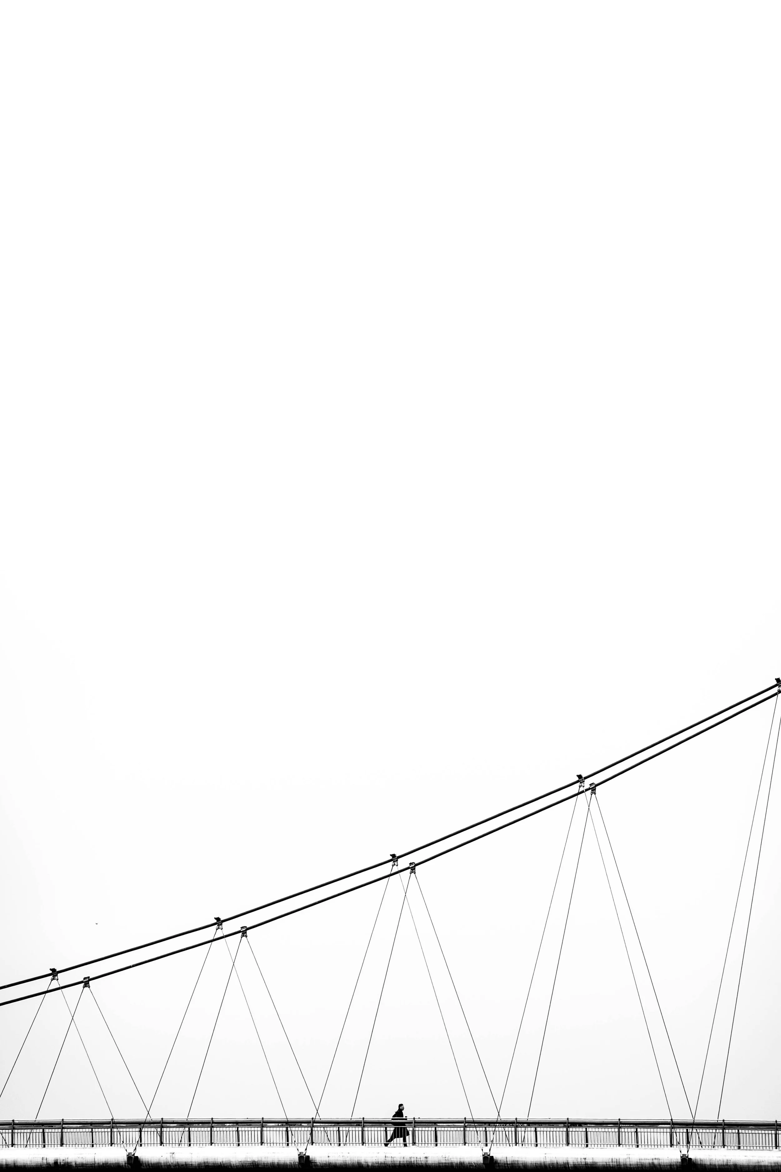 Eine einzelne Person läuft auf einer Brücke mit dicken Stahlseilen, die das Brückengeländer stützen, vor einem weißen Himmel.